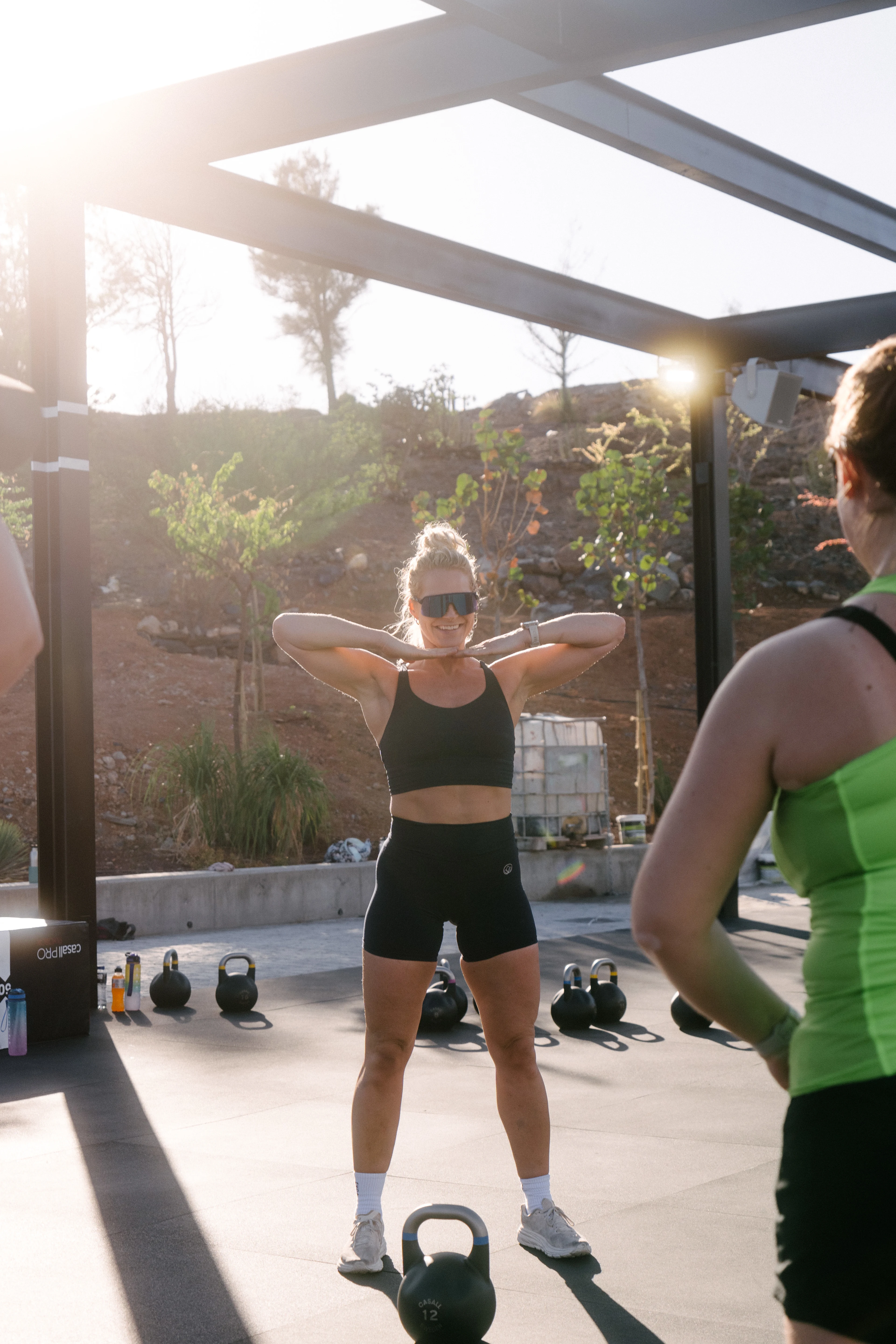 Catrine Collins står med sort topp og treningsshorts samt solbriller og instruerer en dame i gul topp og sort shorts som står med ryggen til.