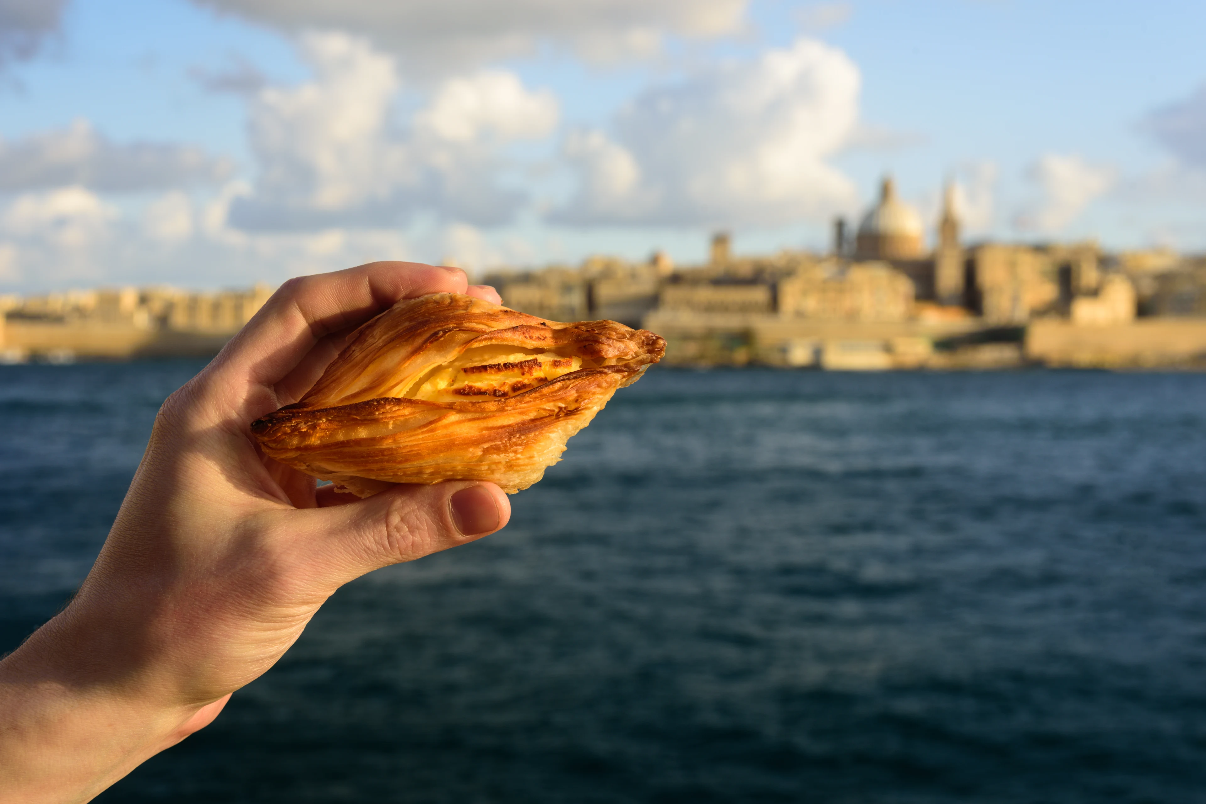 En hånd, der holder en Pastizzi, med udsigt over Valletta.