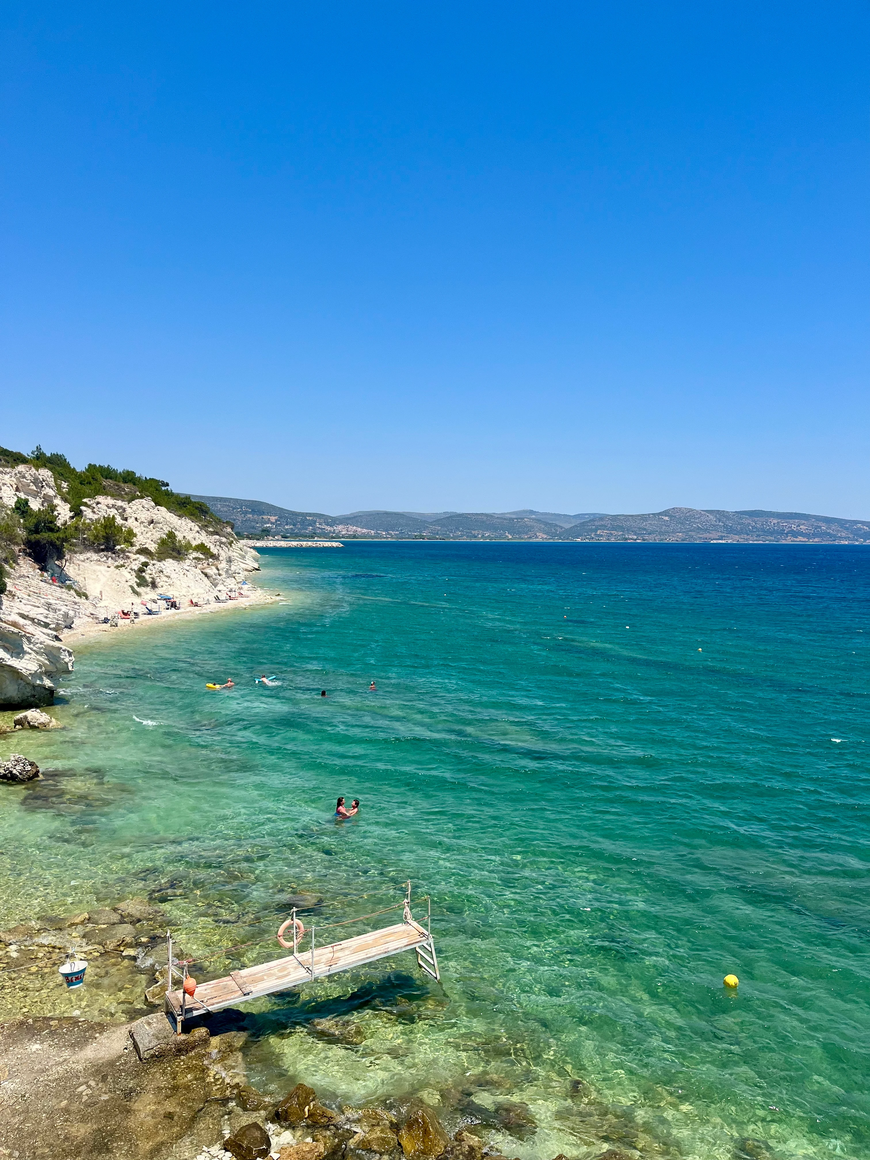 Brygga vid Samos kust med badande människor, klart blått vatten och klippor i bakgrunden – en idyllisk grekisk sommarscen