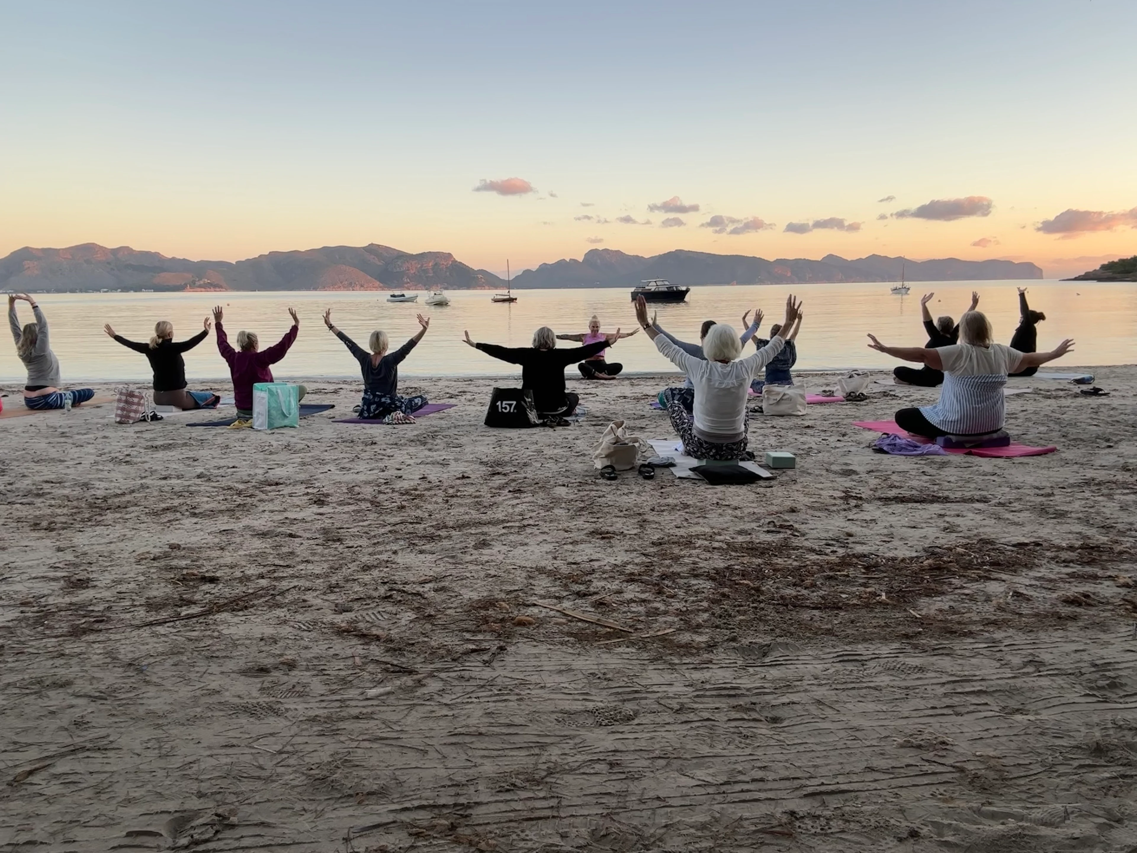 Grupp som deltar i yoga på stranden vid solnedgång, med hav och berg i bakgrunden