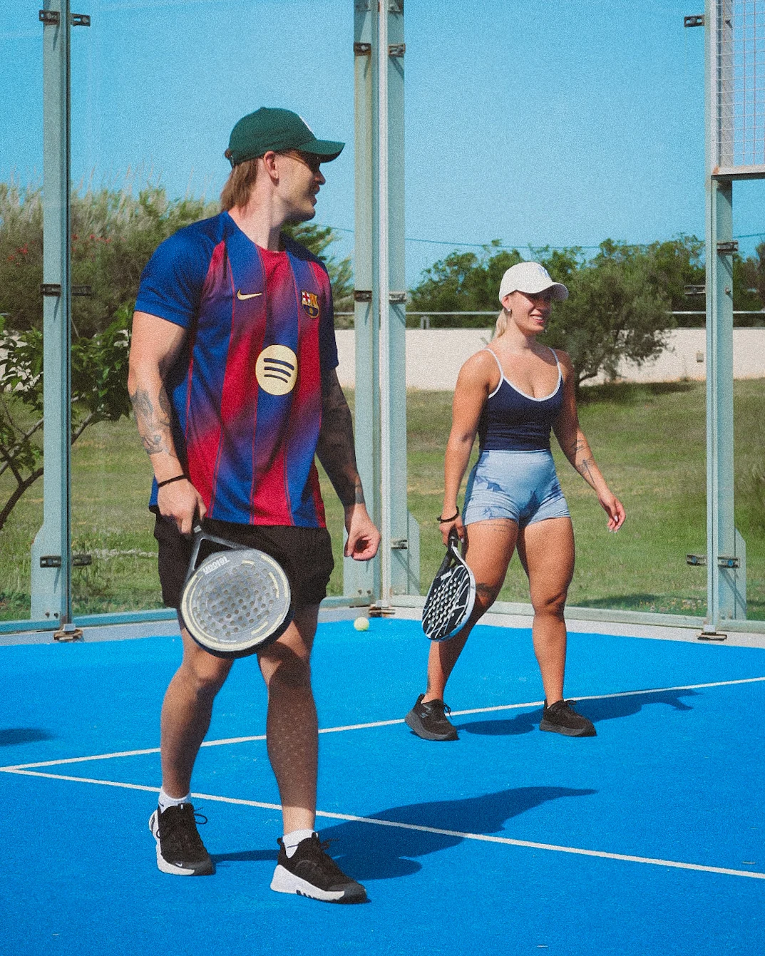 Mies ja nainen aurinkoisella padel-kentällä mailat kädessä