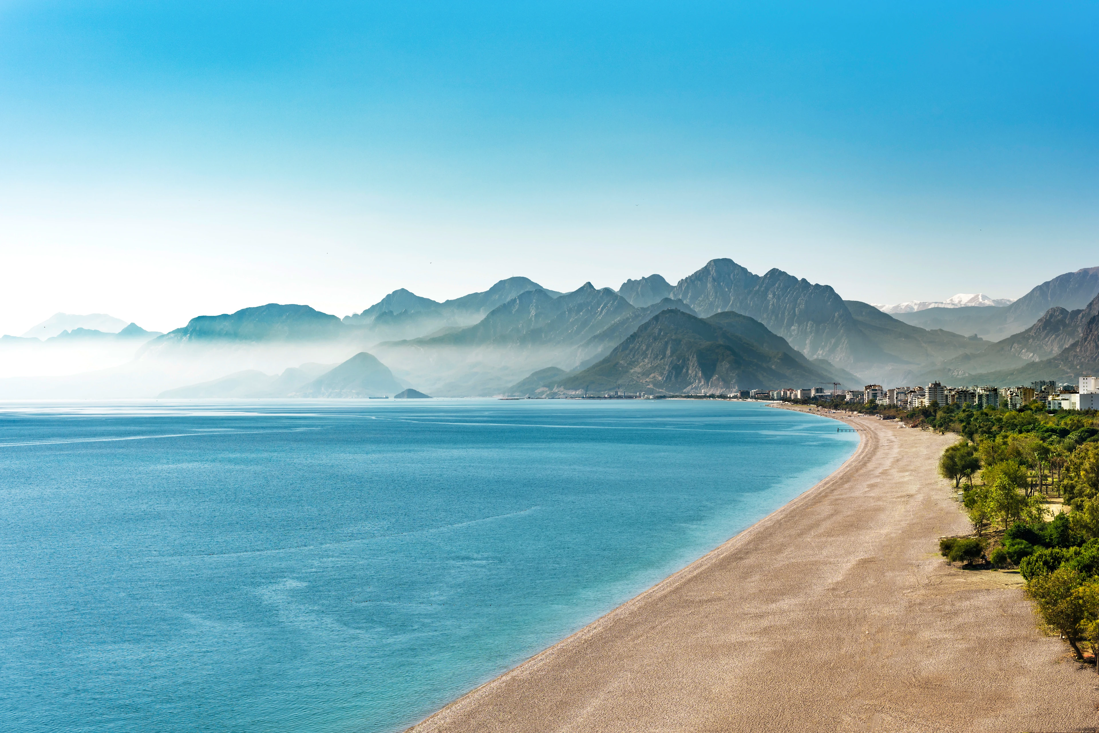 Utsikt over Konyaalti Beach med en lang strandlinje, klart blått hav og en bakgrunn av høye fjell under en klar himmel.