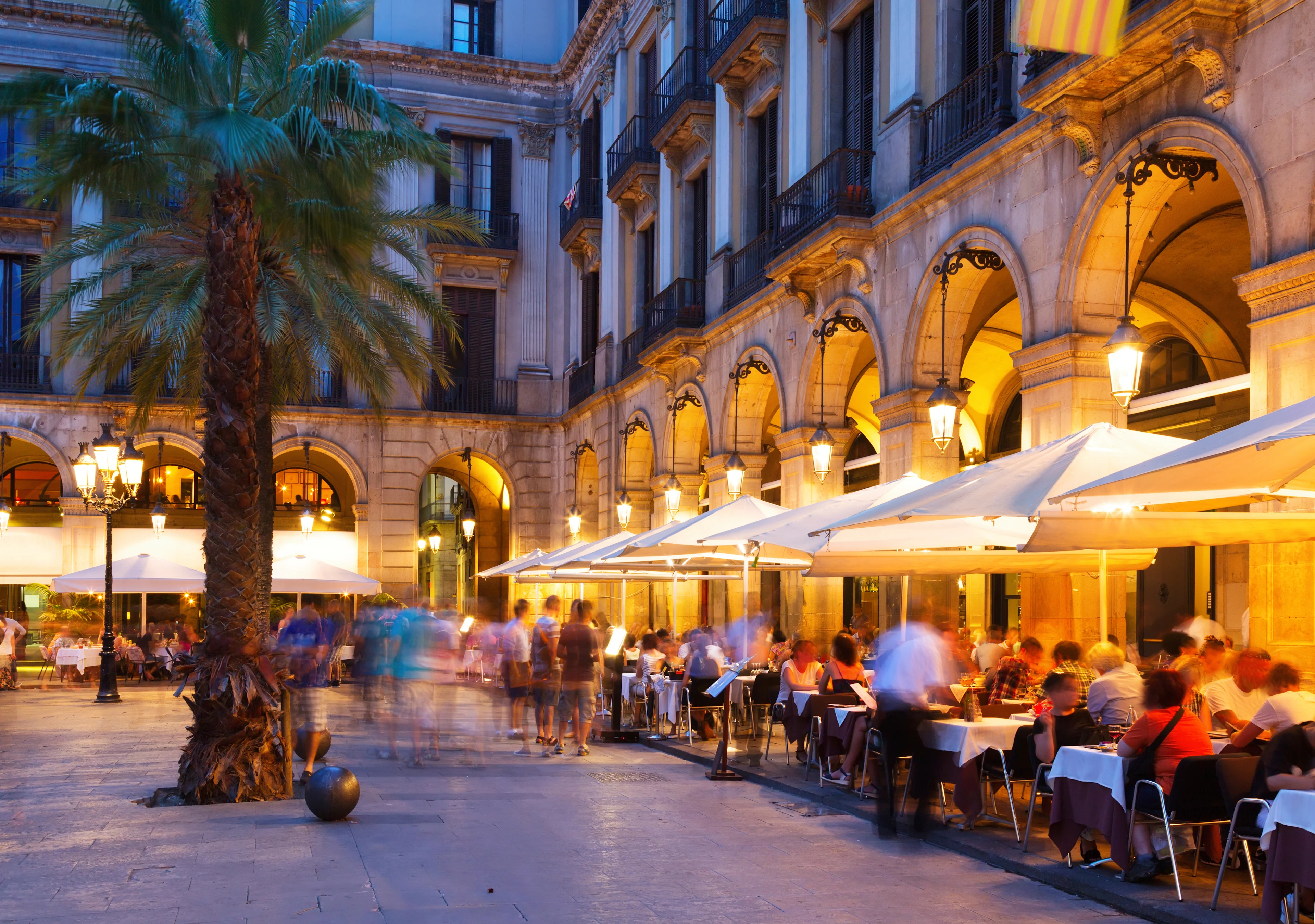 Uteserveringar på Plaça Reial i Barcelona med upplysta arkader och palmer i kvällsljus.