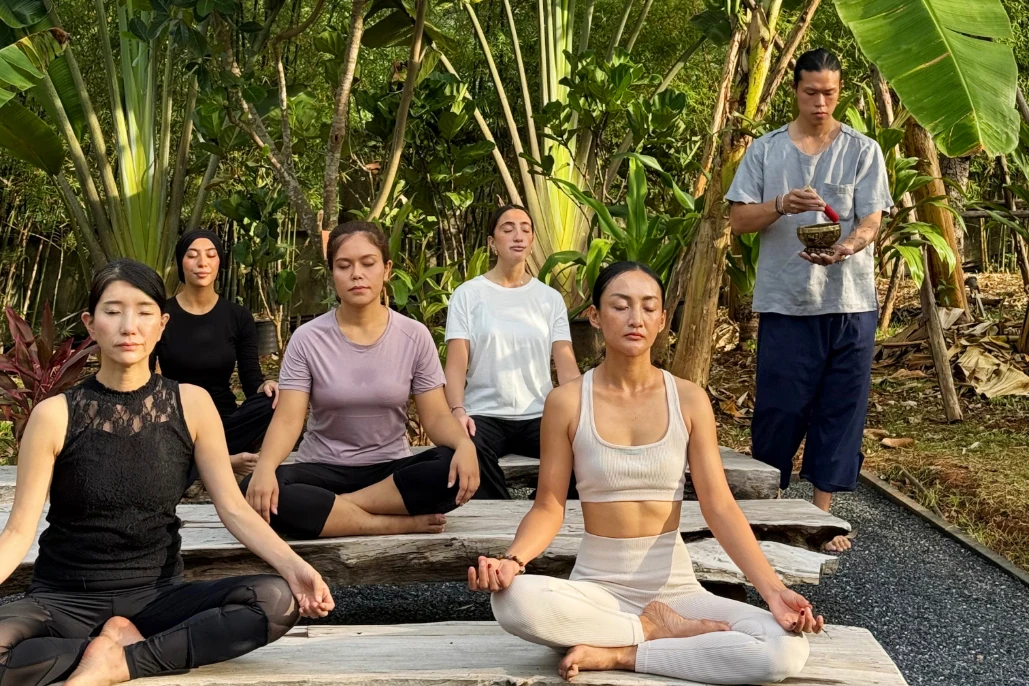 En gruppe personer sidder på yogamåtter og mediterer udendørs i en frodig have, mens en instruktør står i nærheden.
