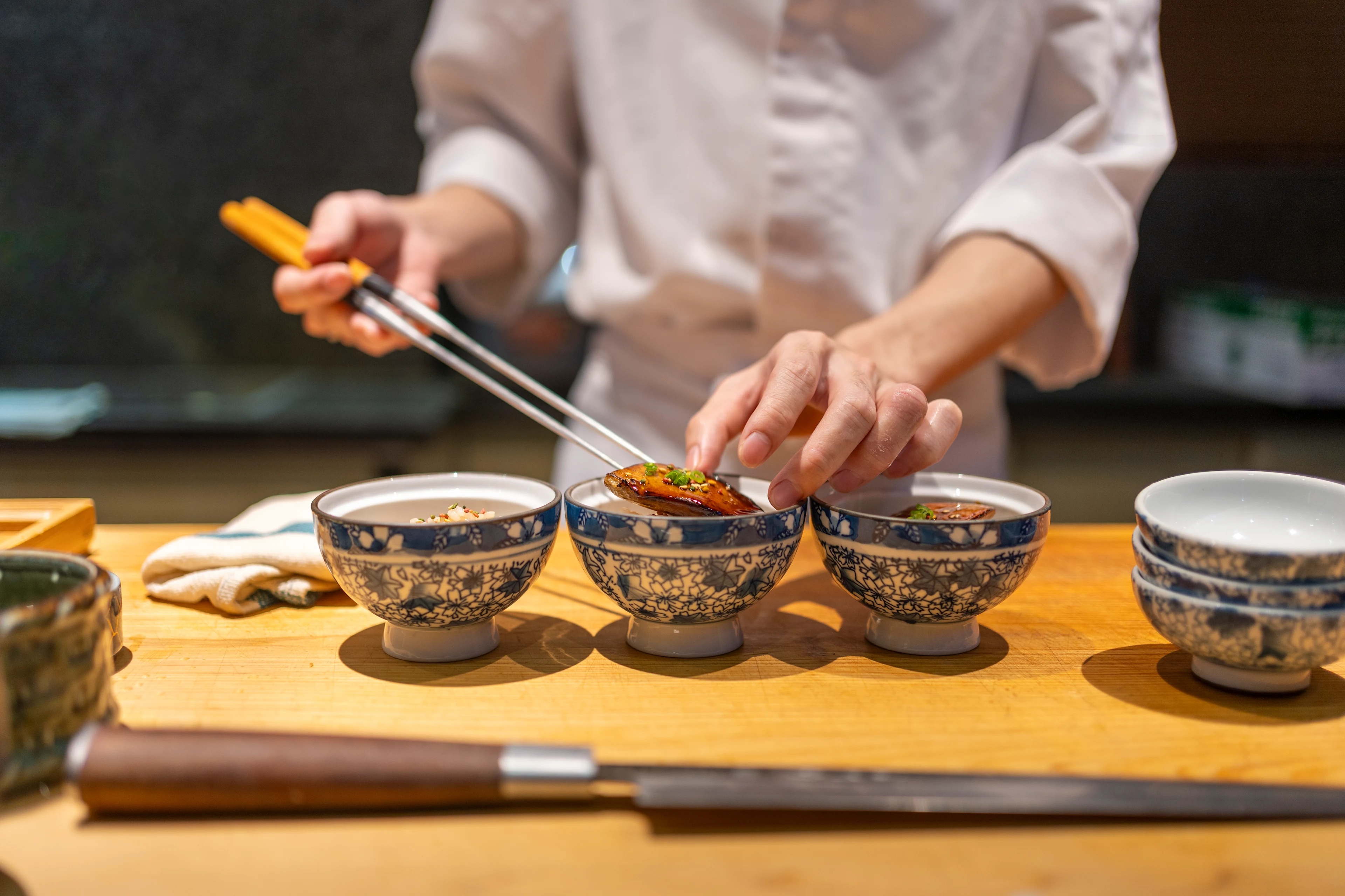 Kok anretter en tilberedt ret mad i mønstrede skåle på en træbænk med japanske spisepinde.
