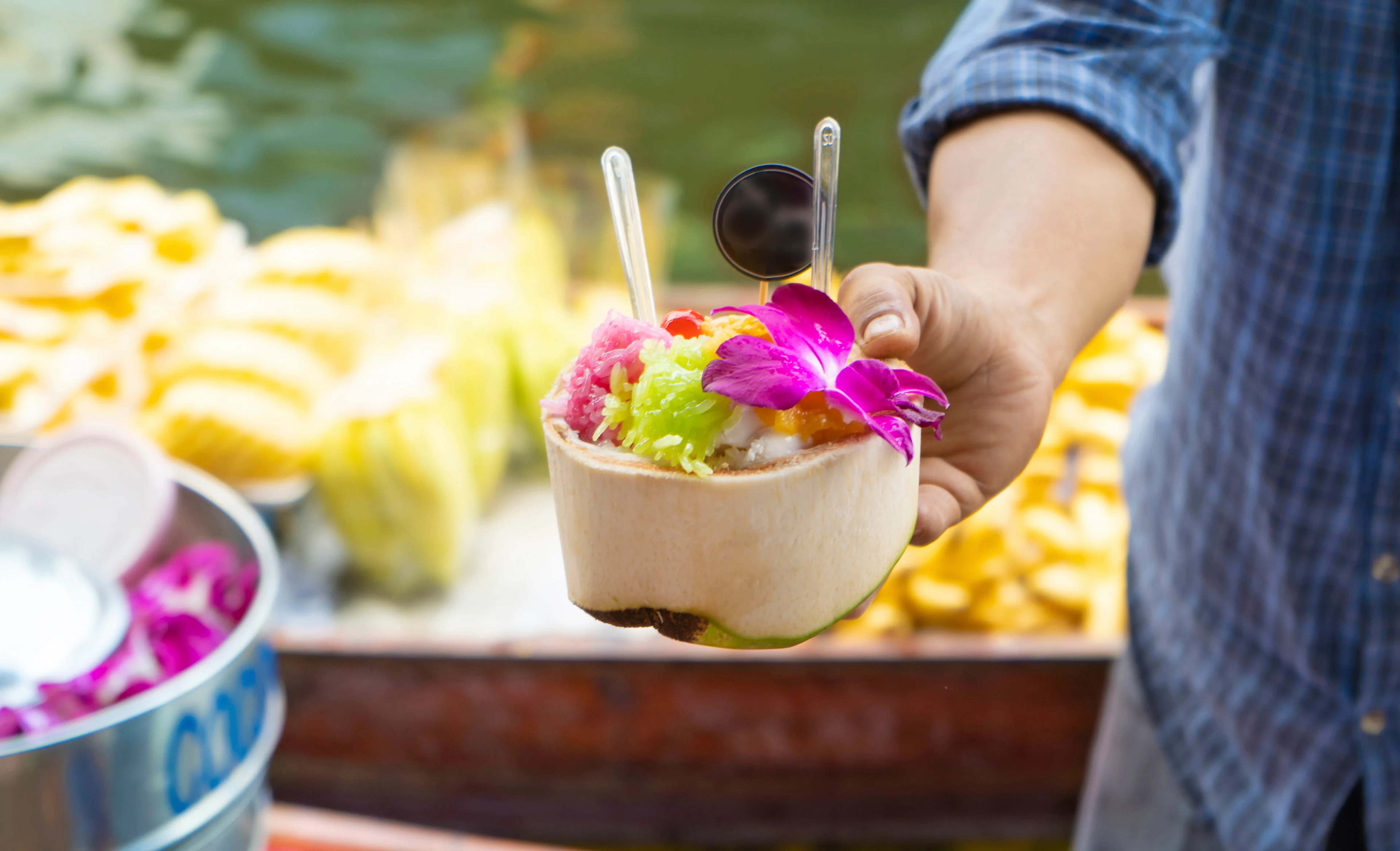 En mann i blårutet skjorte holder en liten skål med fargerik thailandsk dessert toppet med lilla blomst og skje. I bakgrunnen vises et matmarked med fersk frukt og båter langs en kanal i Bangkok