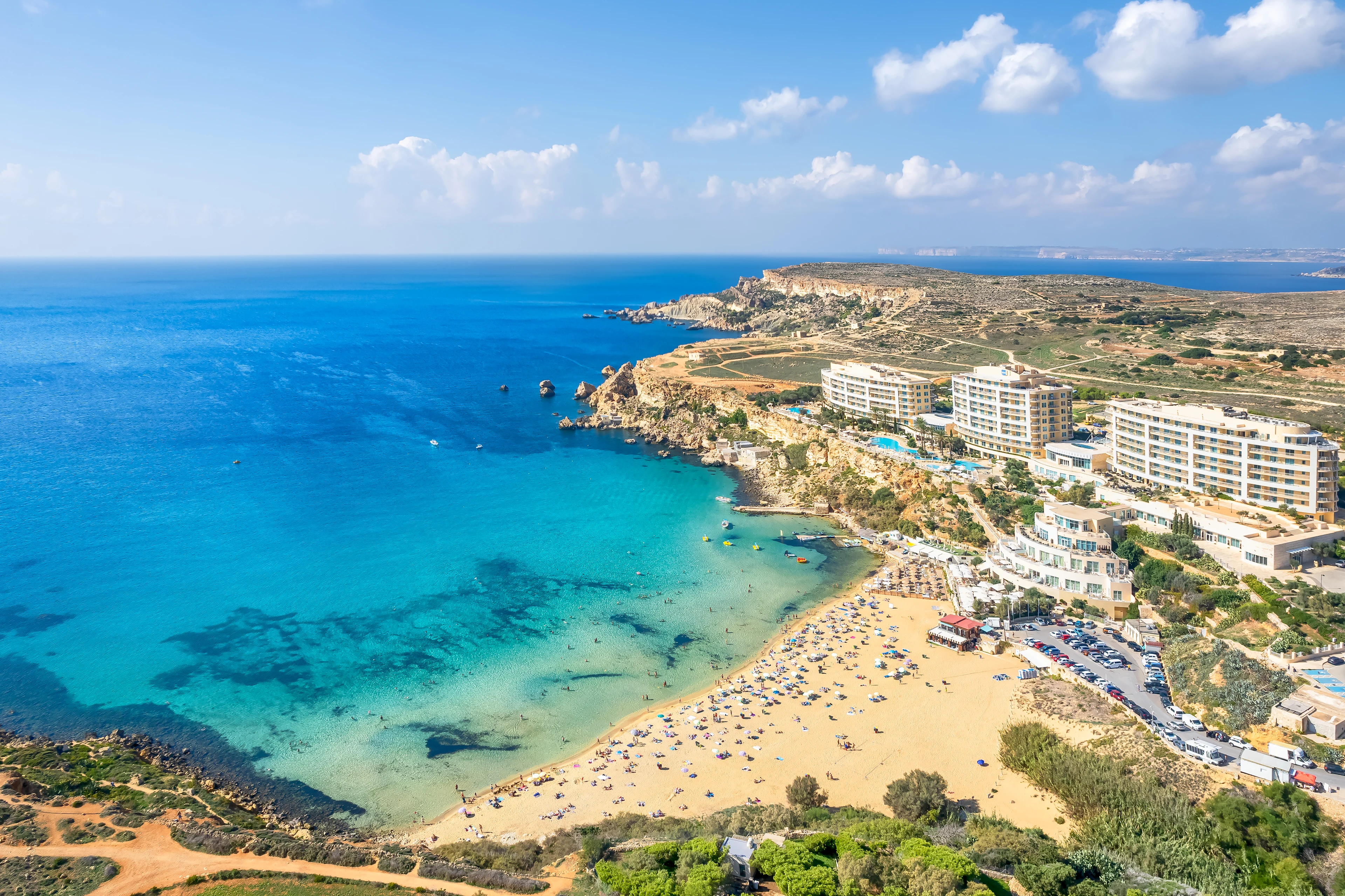 Flygfoto över Golden Bay på Malta med gyllene sandstrand, turkost hav och hotell längs kusten.