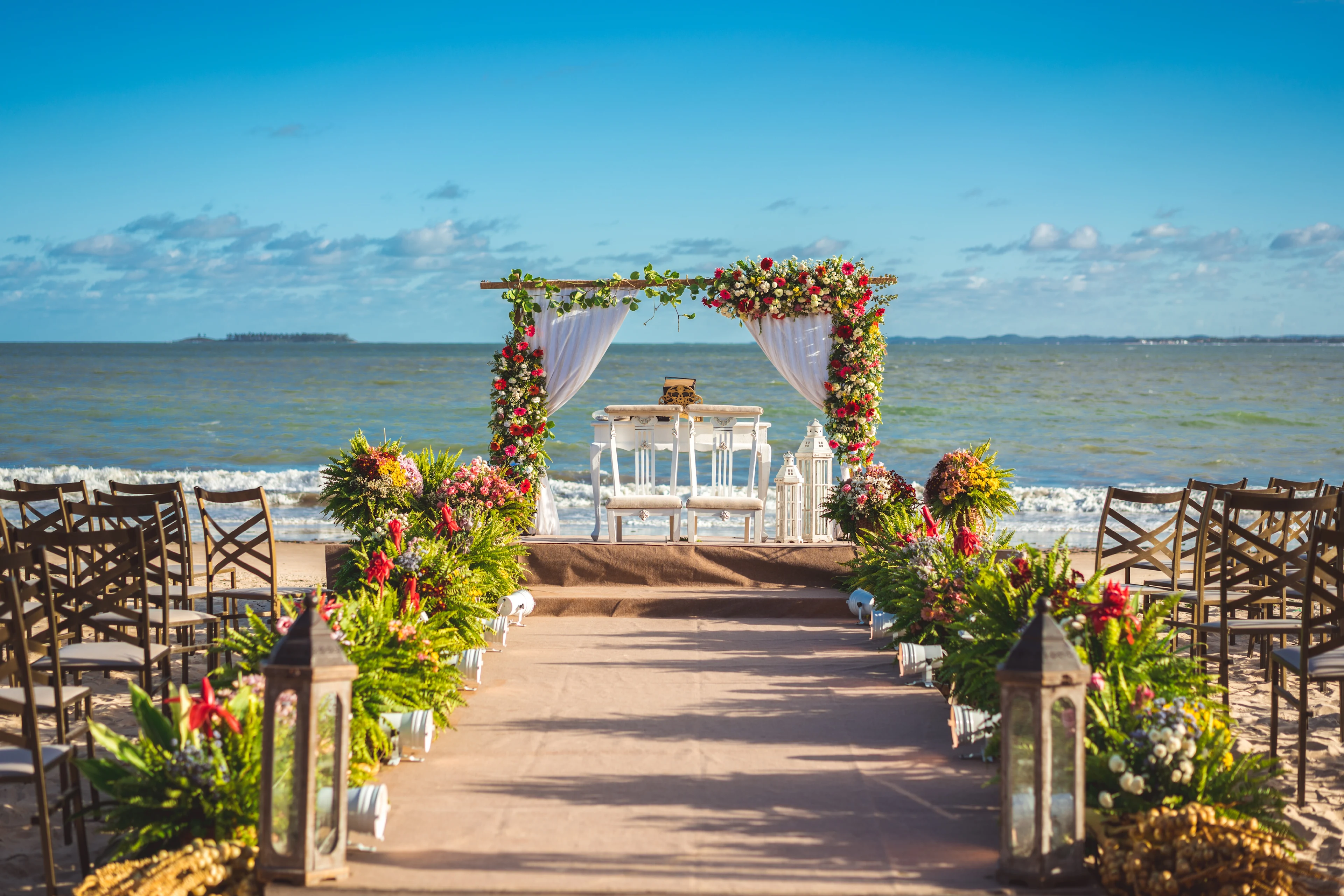 Vakker strandbryllupsseremoni med blomsterdekorert bue, stoler og havet i bakgrunnen under en blå himmel.