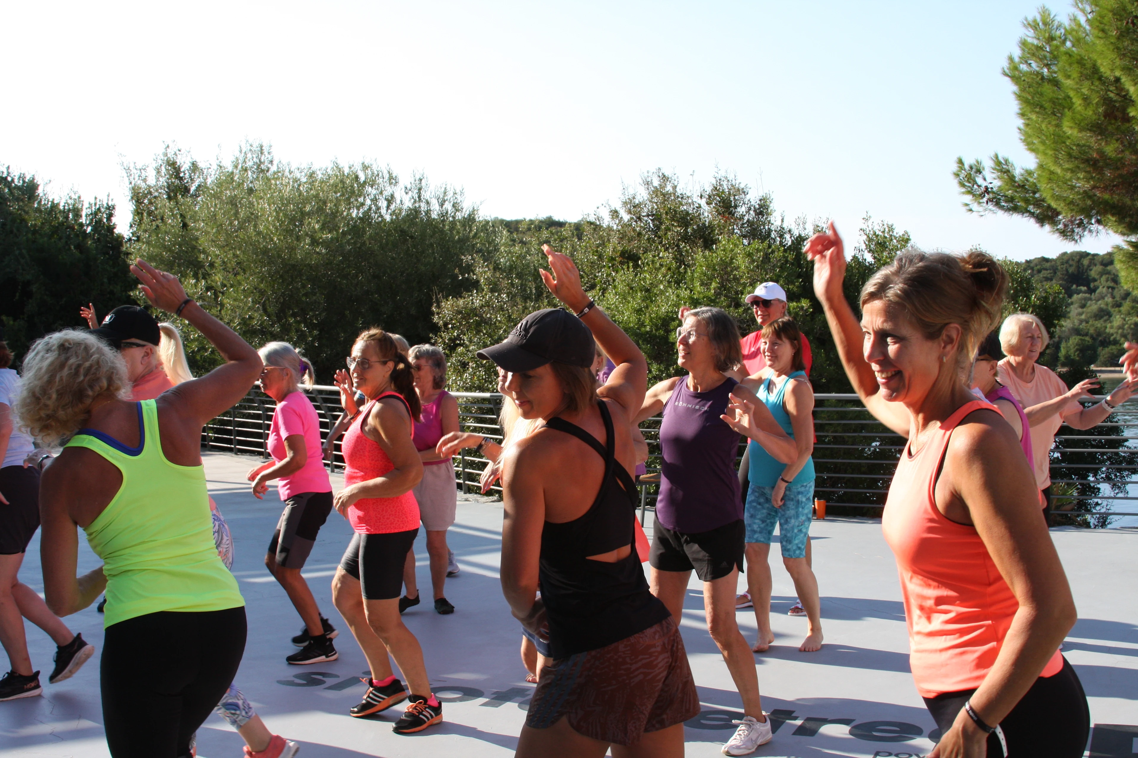 En grupp personer deltar i ett utomhusdanspass på en solig plats omgiven av träd
