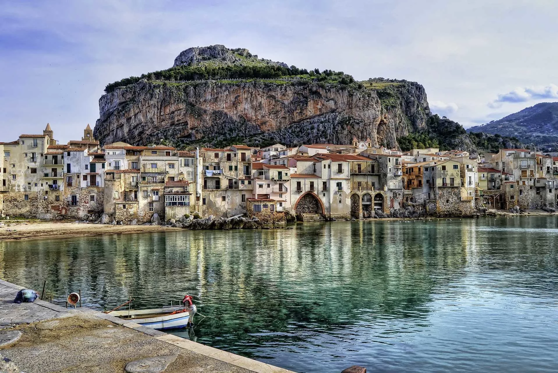 Kystby på Sicilien med historiske bygninger ved havet og en klippe i baggrunden.