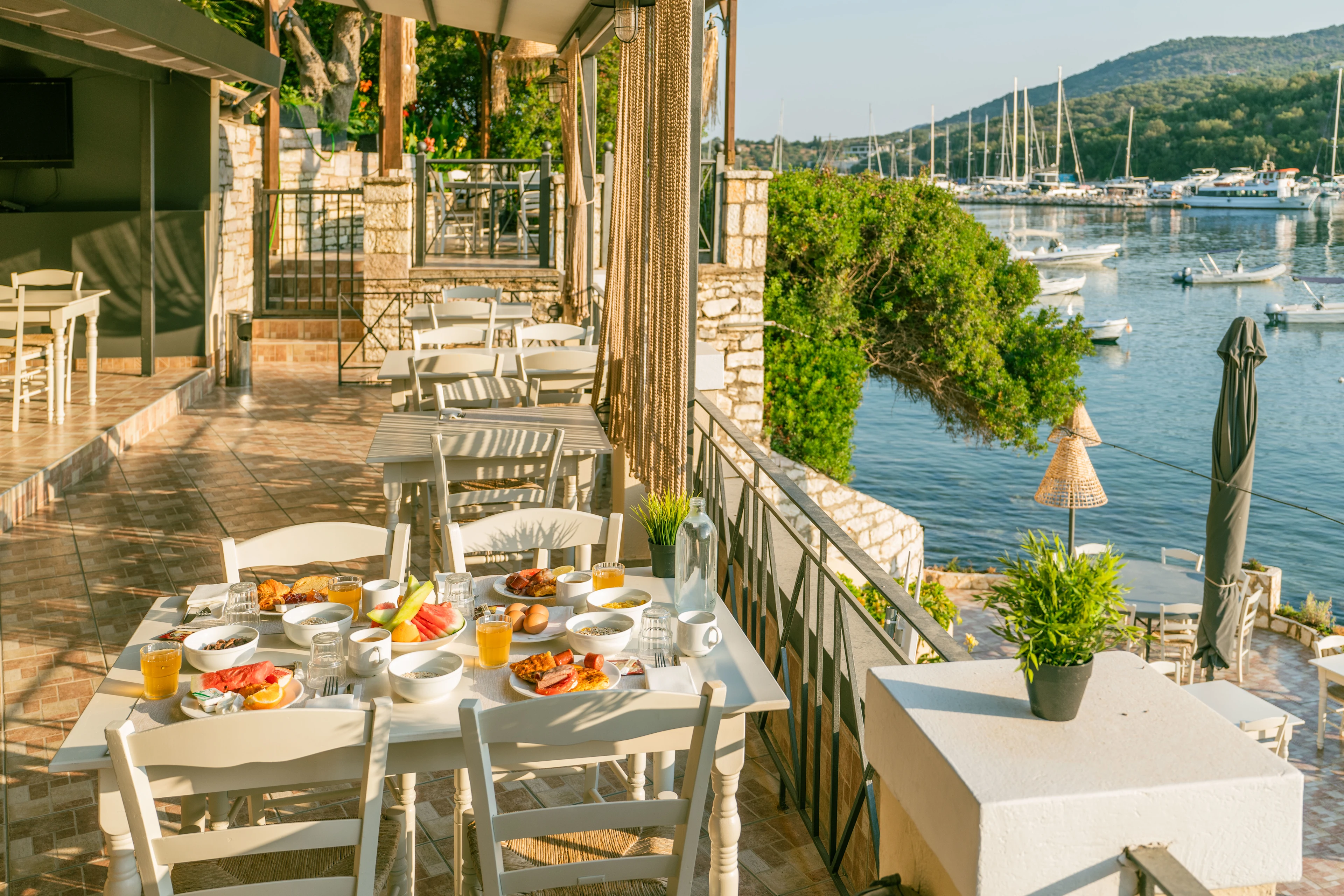 Poolområde med solstole og parasoller på et hotel i Grækenland, omgivet af grønne områder og bjerge i baggrunden.