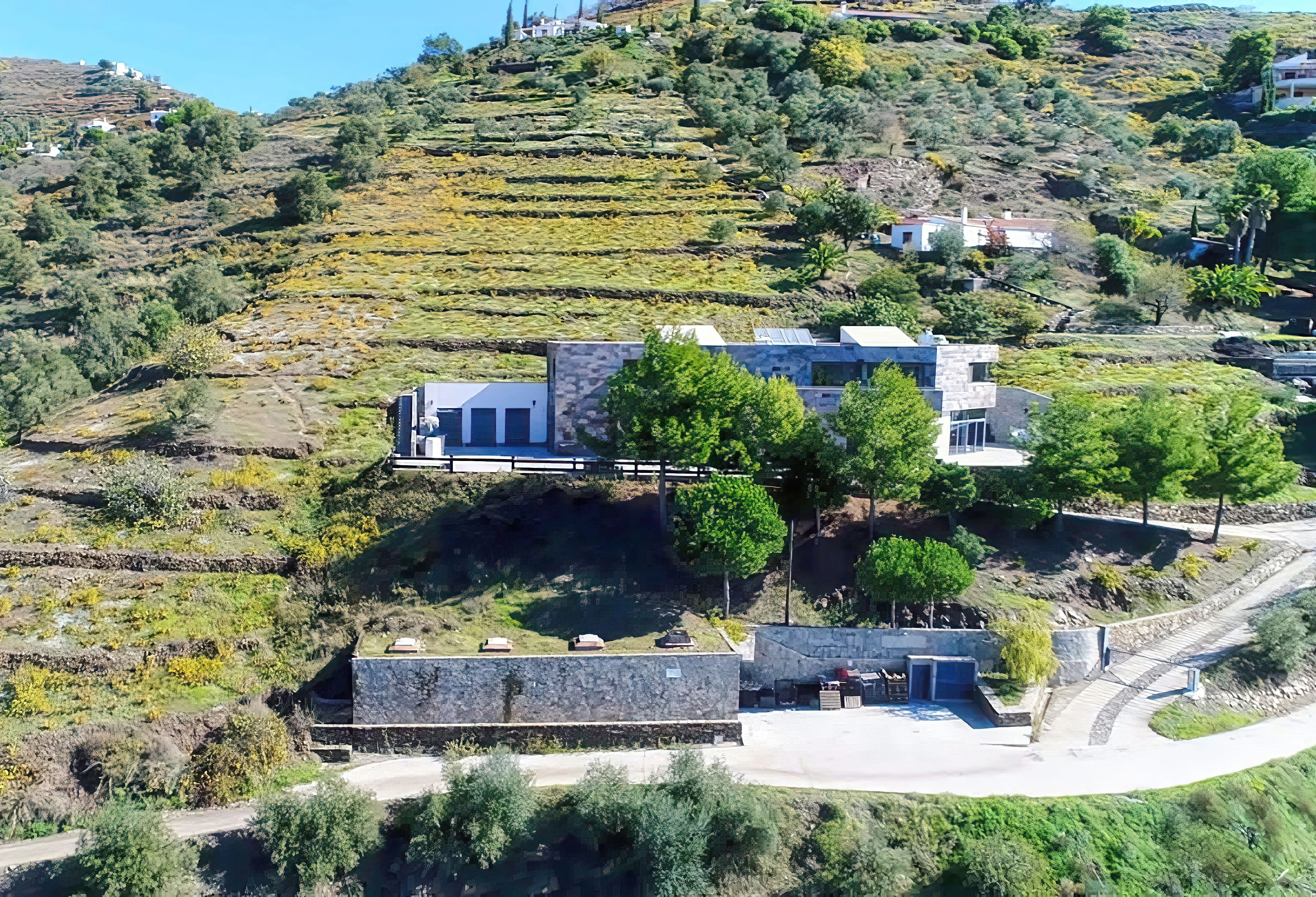 Bodegas Bentomiz omgivet av terrasserade vinodlingar i ett kuperat landskap med modern och äldre arkitektur.