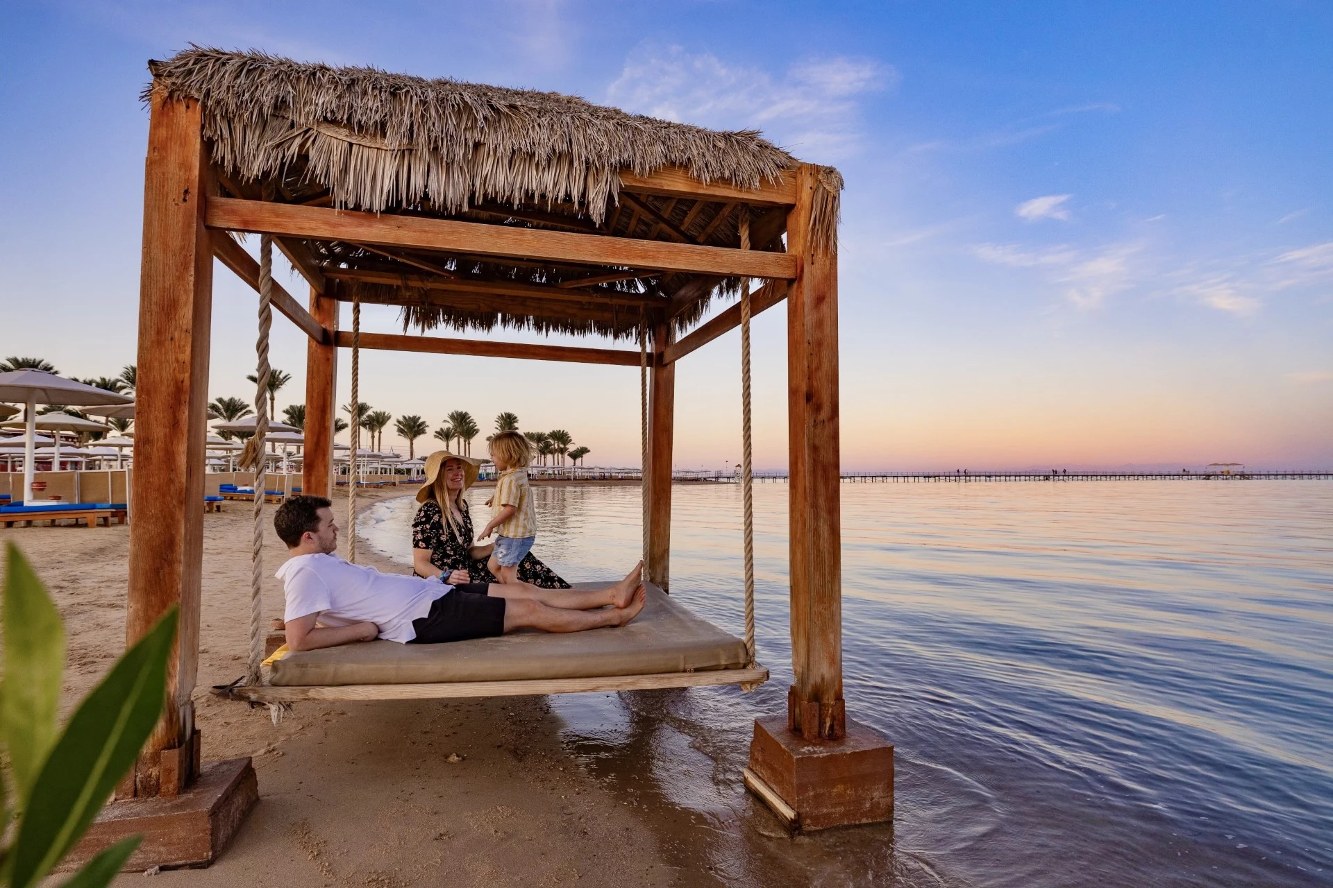 En familj slappnar av på en strandgunga vid vattnet i solnedgången, med palmer och en lång brygga i bakgrunden.