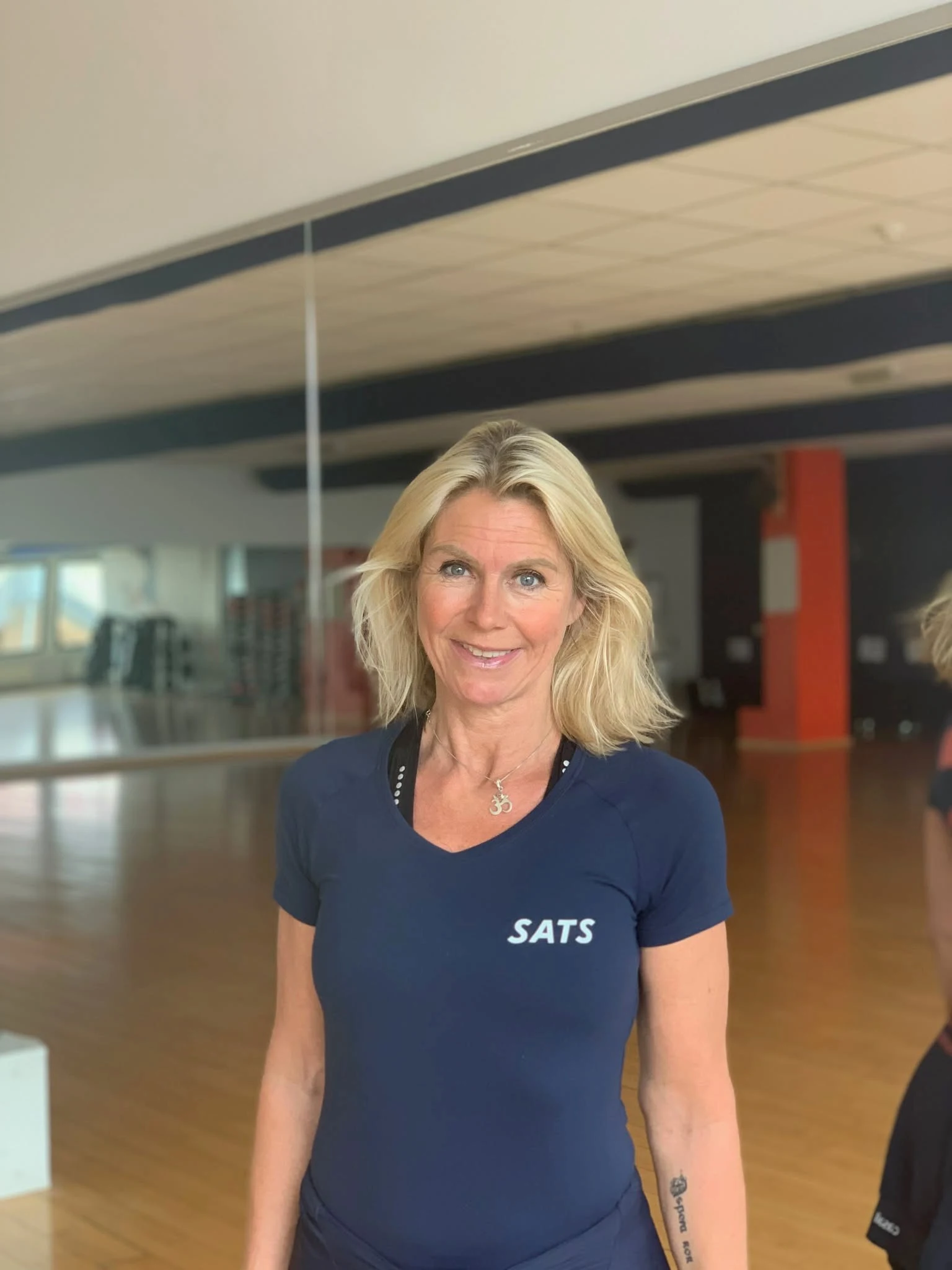 Profilbilde av Trine Tschudi med blå t-skjorte med påskriften SATS, der hun står i en treningssal