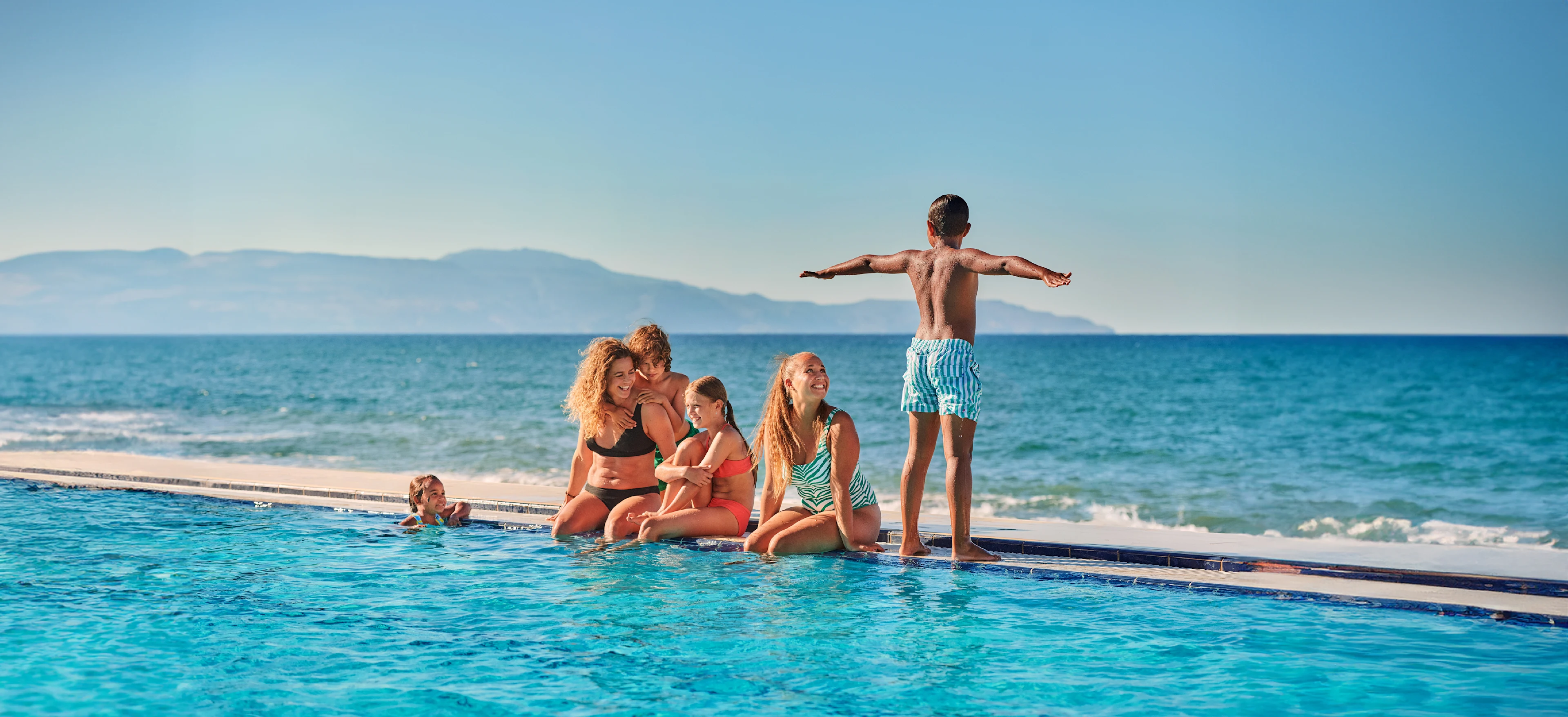 Børn og voksne hygger sig ved en pool ved stranden med udsigt til havet og bjerge i baggrunden. Solrigt og sommerligt.