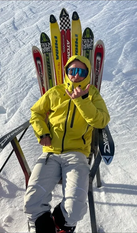 Person sidder i alperne i gul skijakke i en lænestol bygget af gamle ski i solskin.