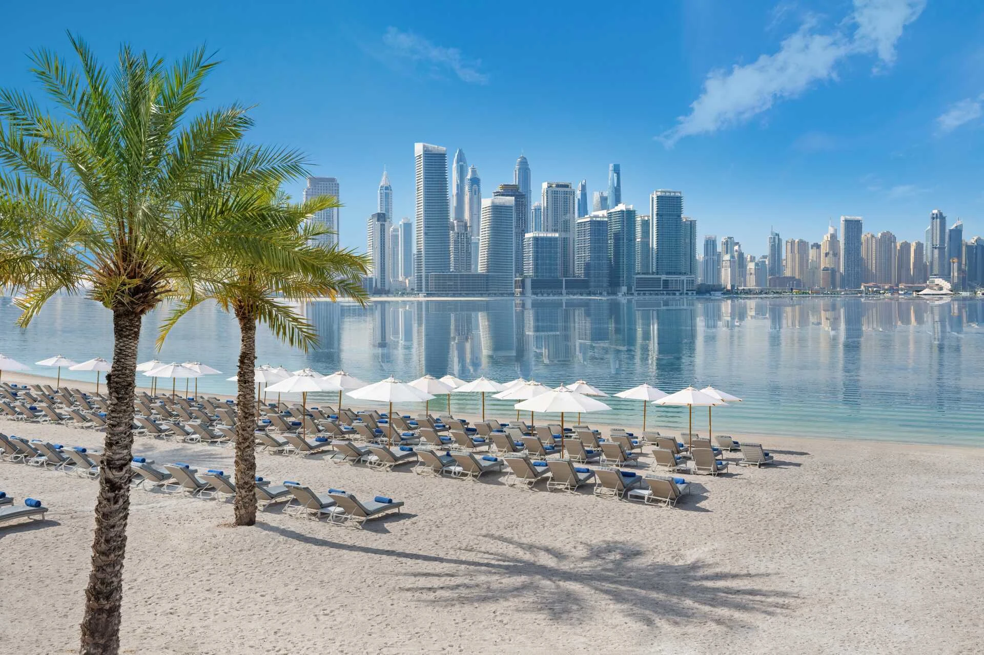 Hiekkaranta palmujen ja kävelyteiden kanssa, taustalla kaupungin pilvenpiirtäjiä kirkkaan sinisen taivaan alla.