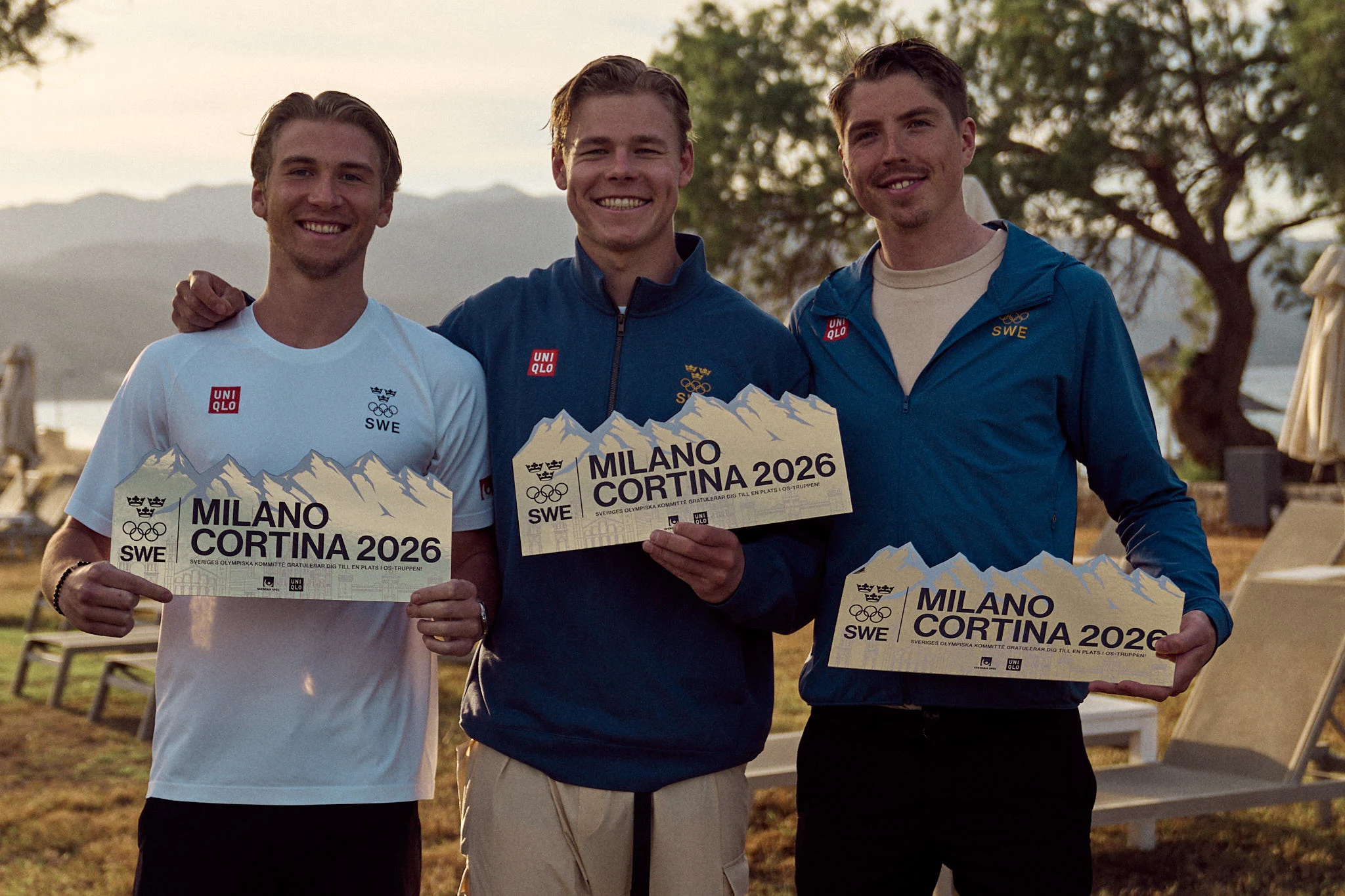 Tre svenska idrottare står utomhus och håller skyltar med texten ”Milano Cortina 2026"