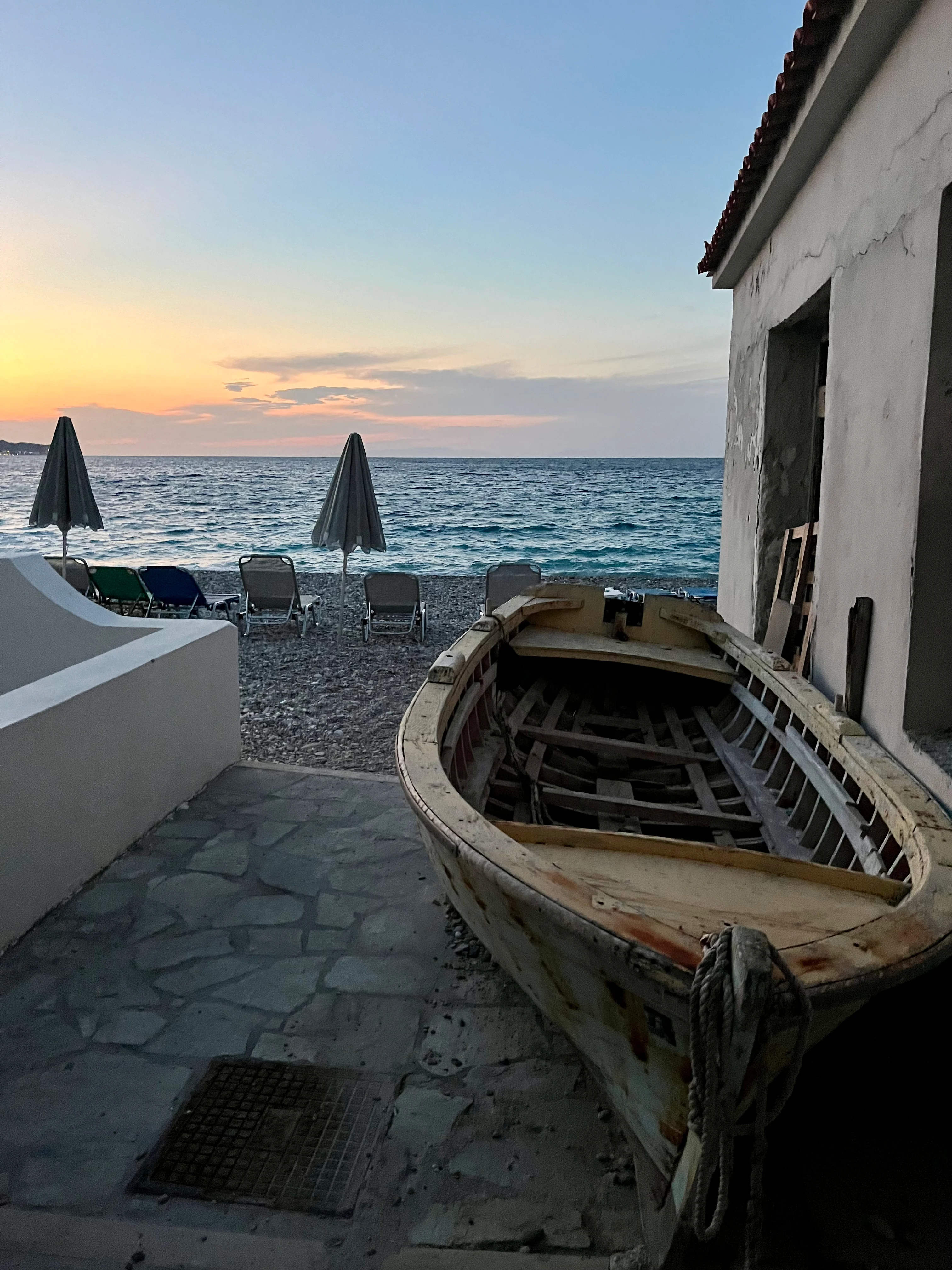 Solnedgång vid stenstrand med gammal träbåt, solstolar och havsutsikt – en stillsam och vacker kustscen på Samos.