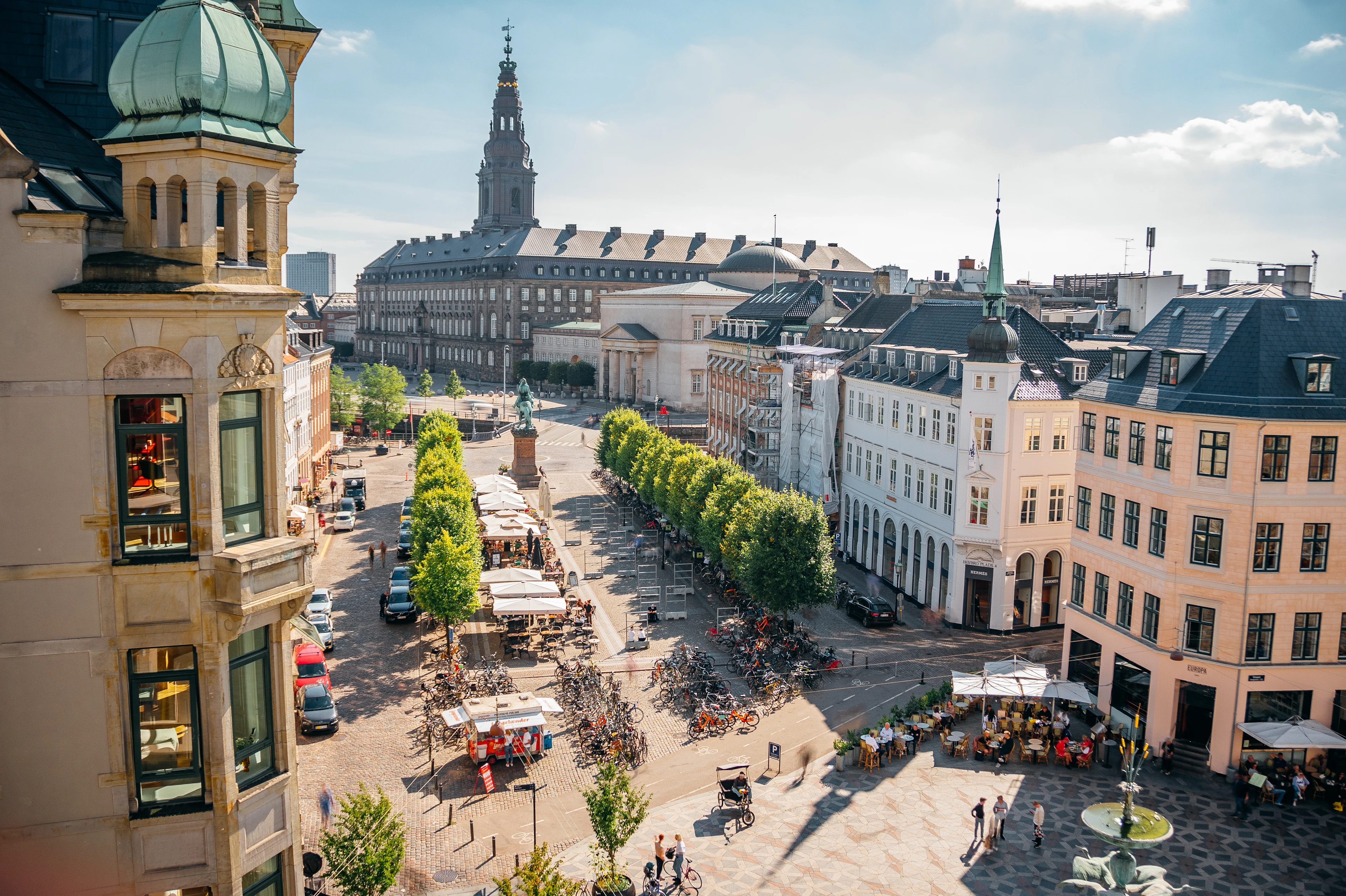 Vy över centrala Köpenhamn med eleganta byggnader, grönskande träd och ett livligt torg fyllt av människor och uteserveringar.