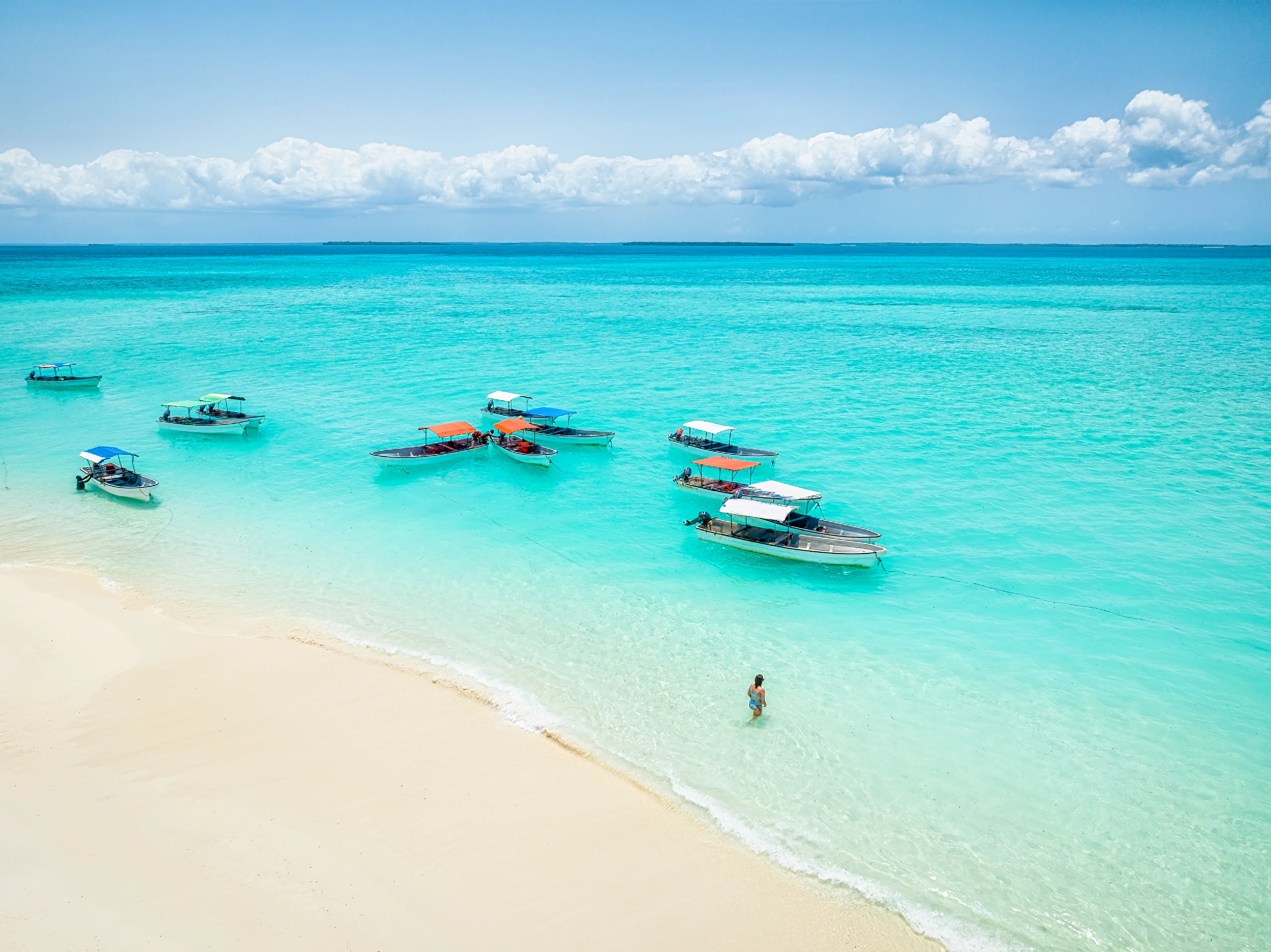 Flygfoto över båtar vid en tropisk strand med vit sand och turkost vatten på Zanzibar. En turist går i det grunda vattnet.