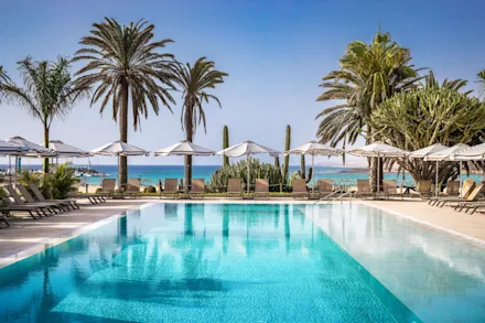 Vackert poolområde med solstolar på ett hotell vid stranden. I bakgrunden ser man det turkosa vattnet på ett strandnära hotell i Gran Canaria.