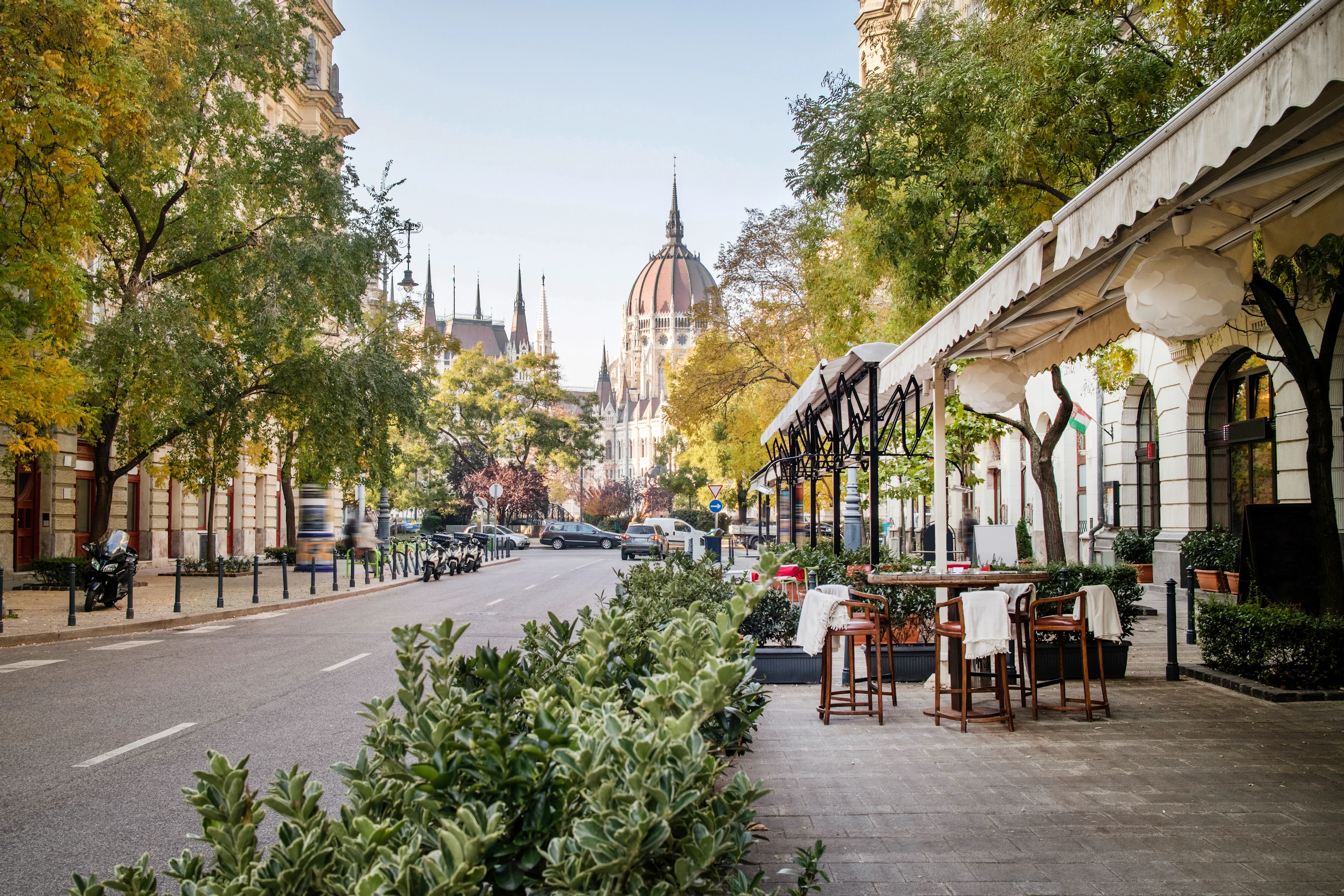 En lugn stadsgata i Budapest kantad av gröna träd och buskar, med uteserveringar till höger och en imponerande byggnad med kupol och spiror i bakgrunden.