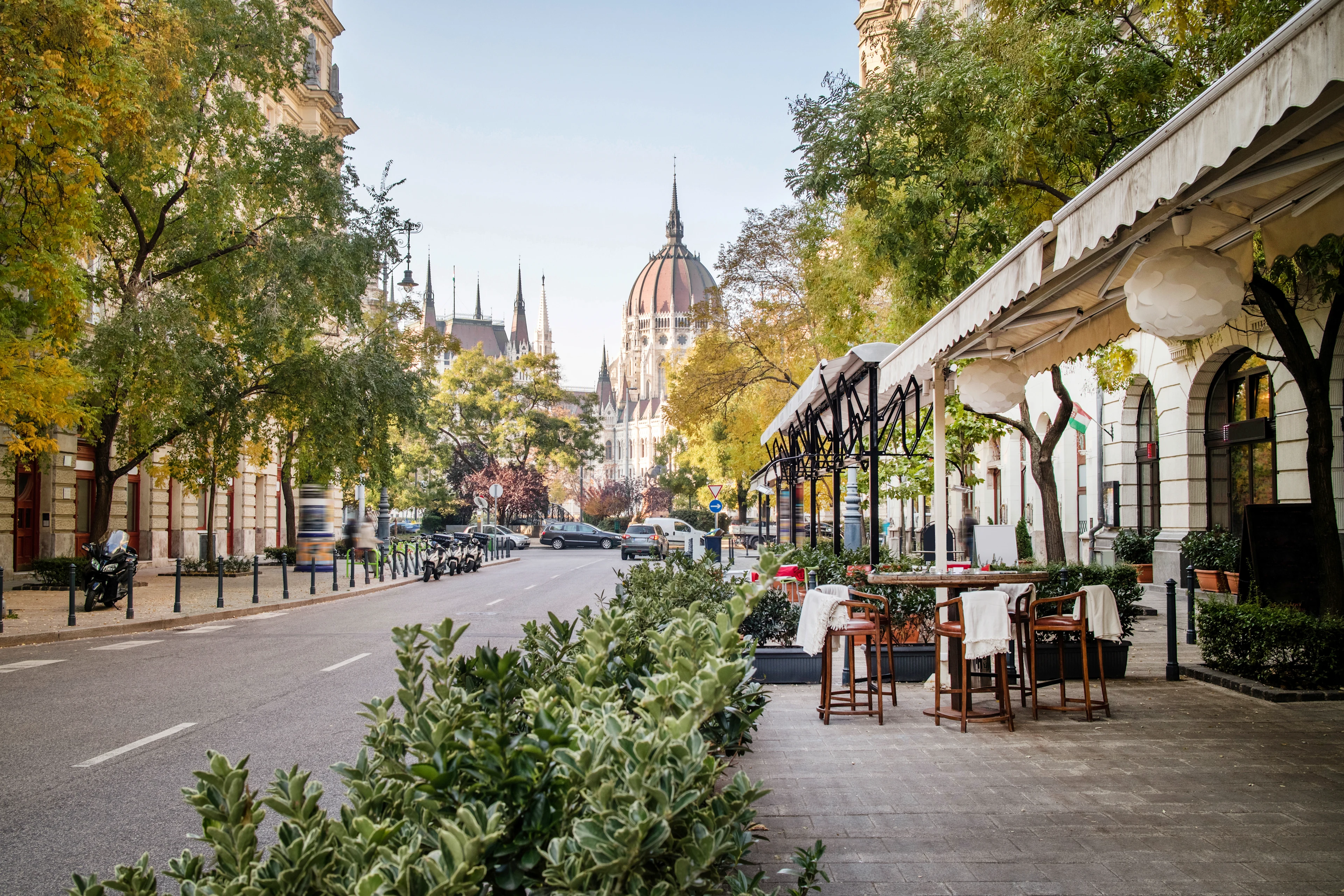 En rolig bygate i Budapest omkranset av grønne trær og busker, med uteservering til høyre og en imponerende bygning med kuppel og spir i bakgrunnen.