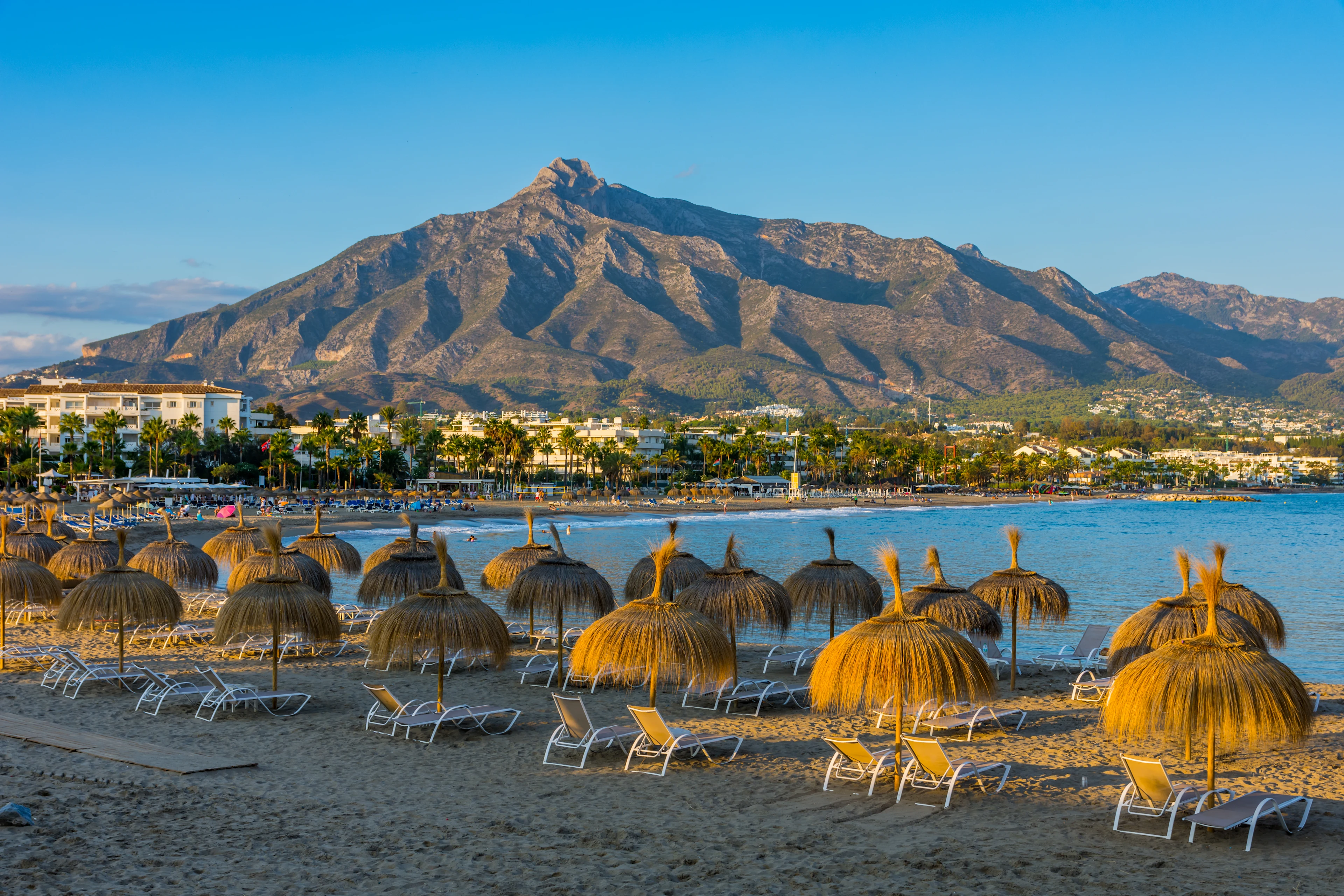 Strand i Puerto Banús med halmparasoller och berg i bakgrunden.
