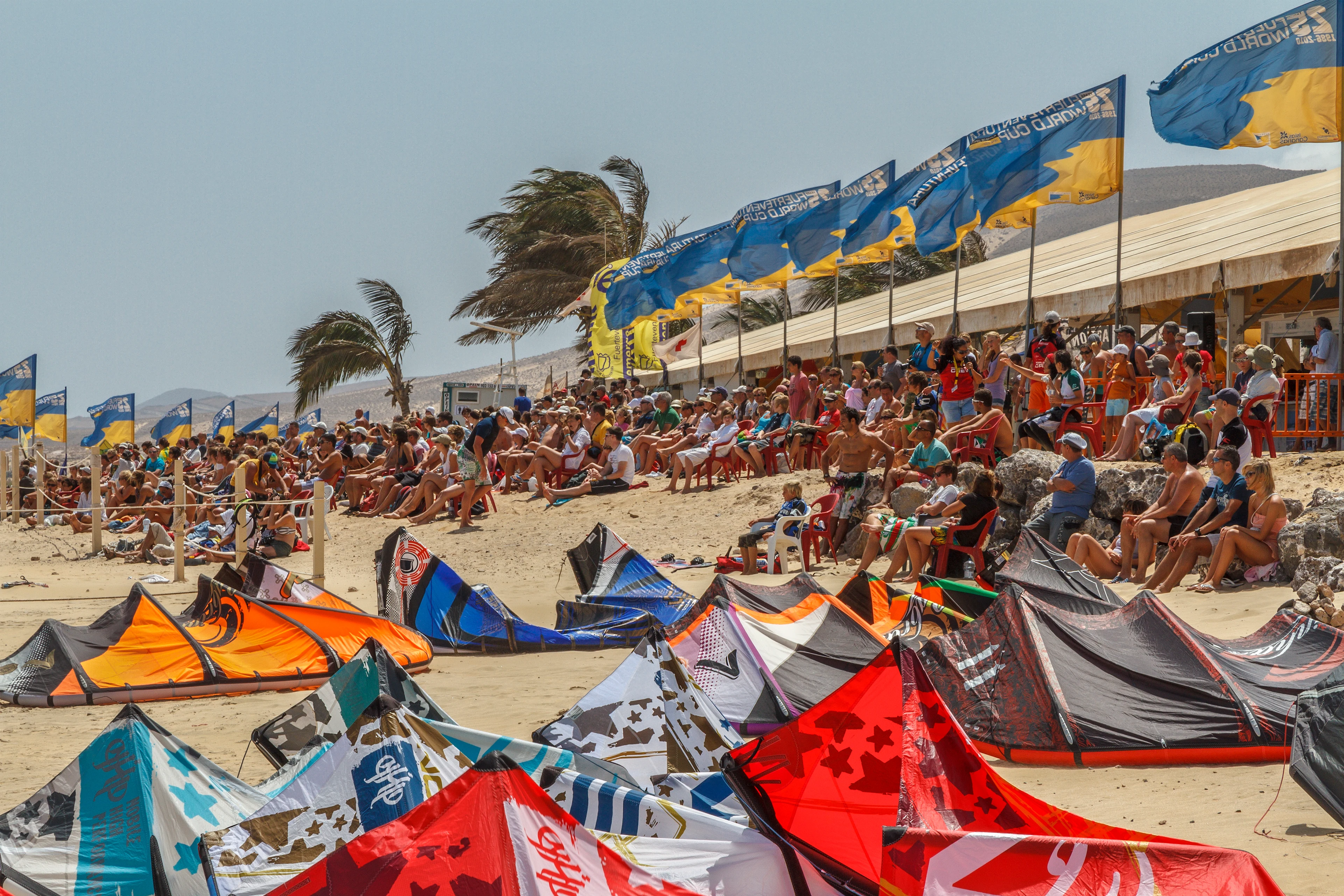 Publikum på strandtribuner på et wind- og vannsportsarrangement, med fargerike drager i sanden og flagg i vinden
