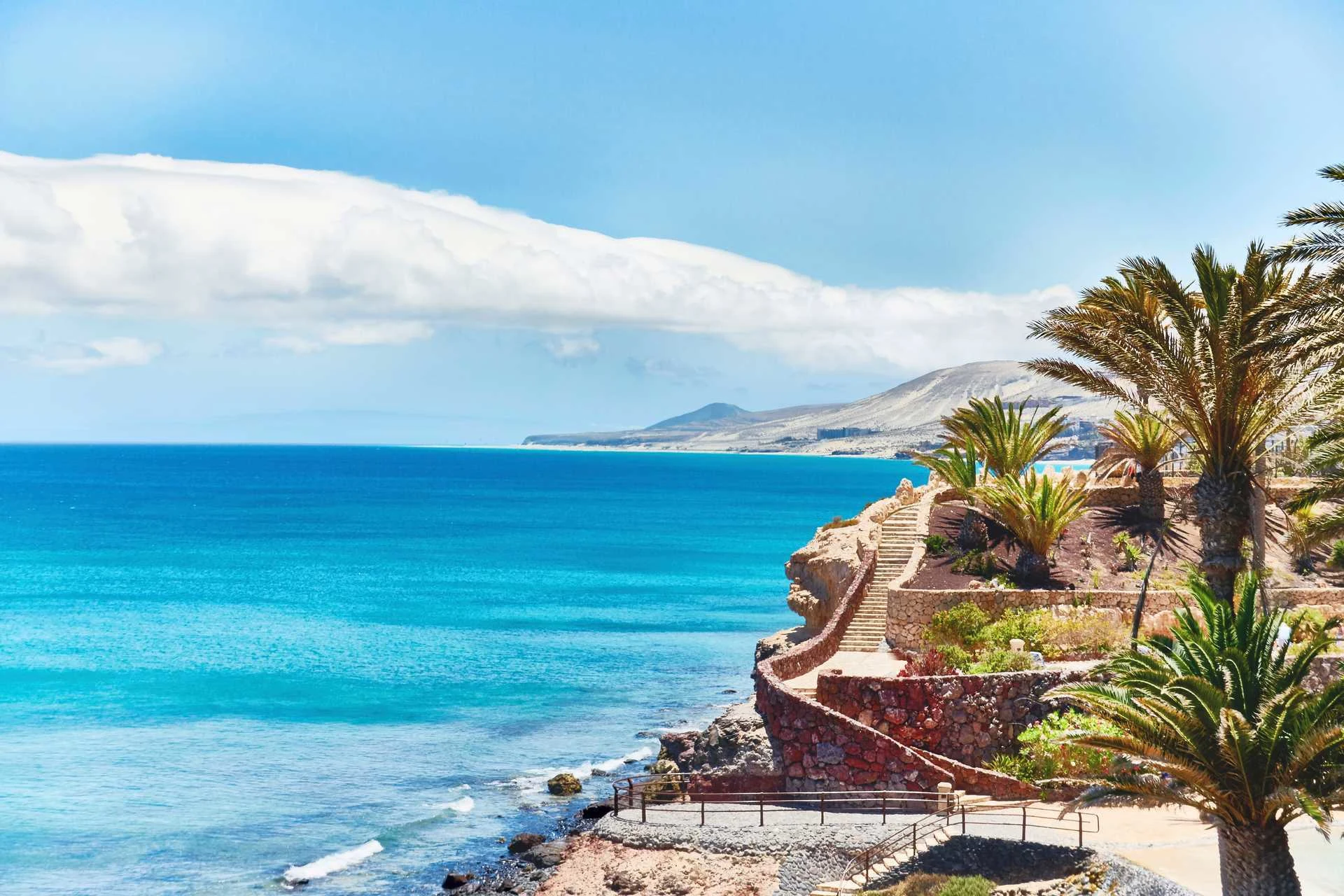 Flott gangsti bygd i stein langs det knallblå havet på Fuerteventura. Palmer på siden og langs gangstien. 