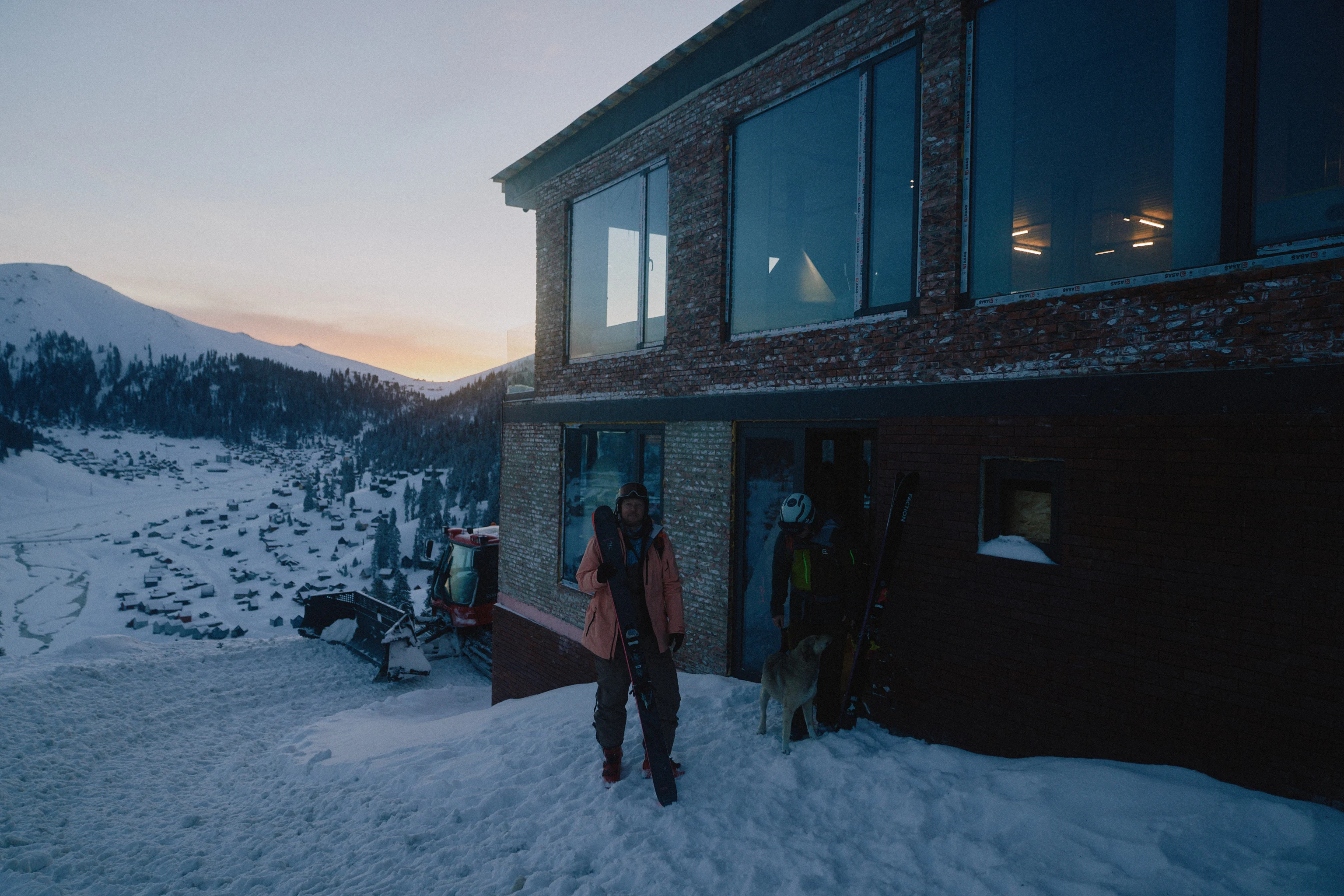 Personer med skidor står utanför en fjällstuga i snötäckt bergsmiljö vid gryning eller skymning, med utsikt över en dal och snöklädda sluttningar.