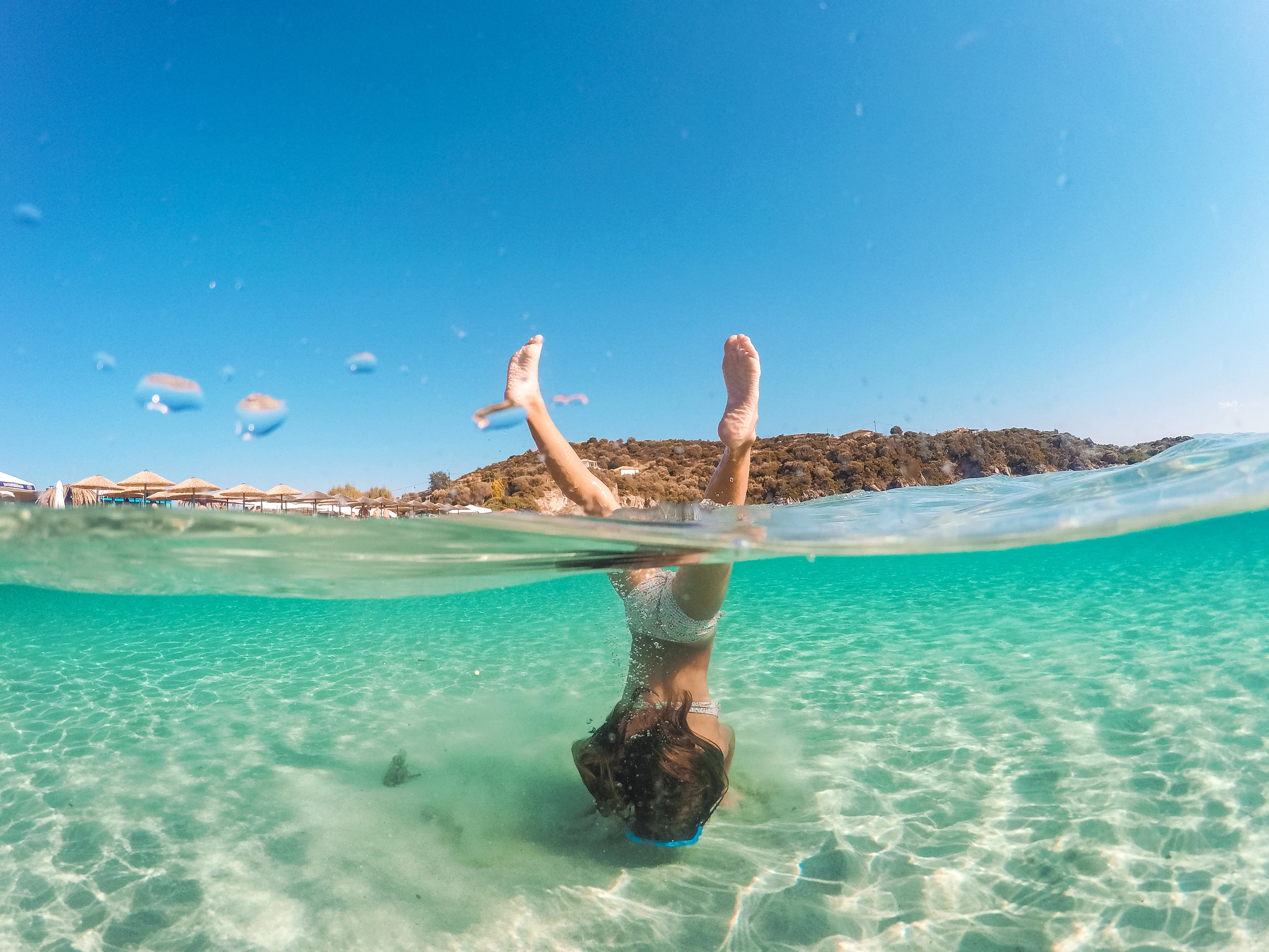 Jente gjør en salto i krystallklart vann på en strand med parasoller i bakgrunnen, sett delvis under vann.