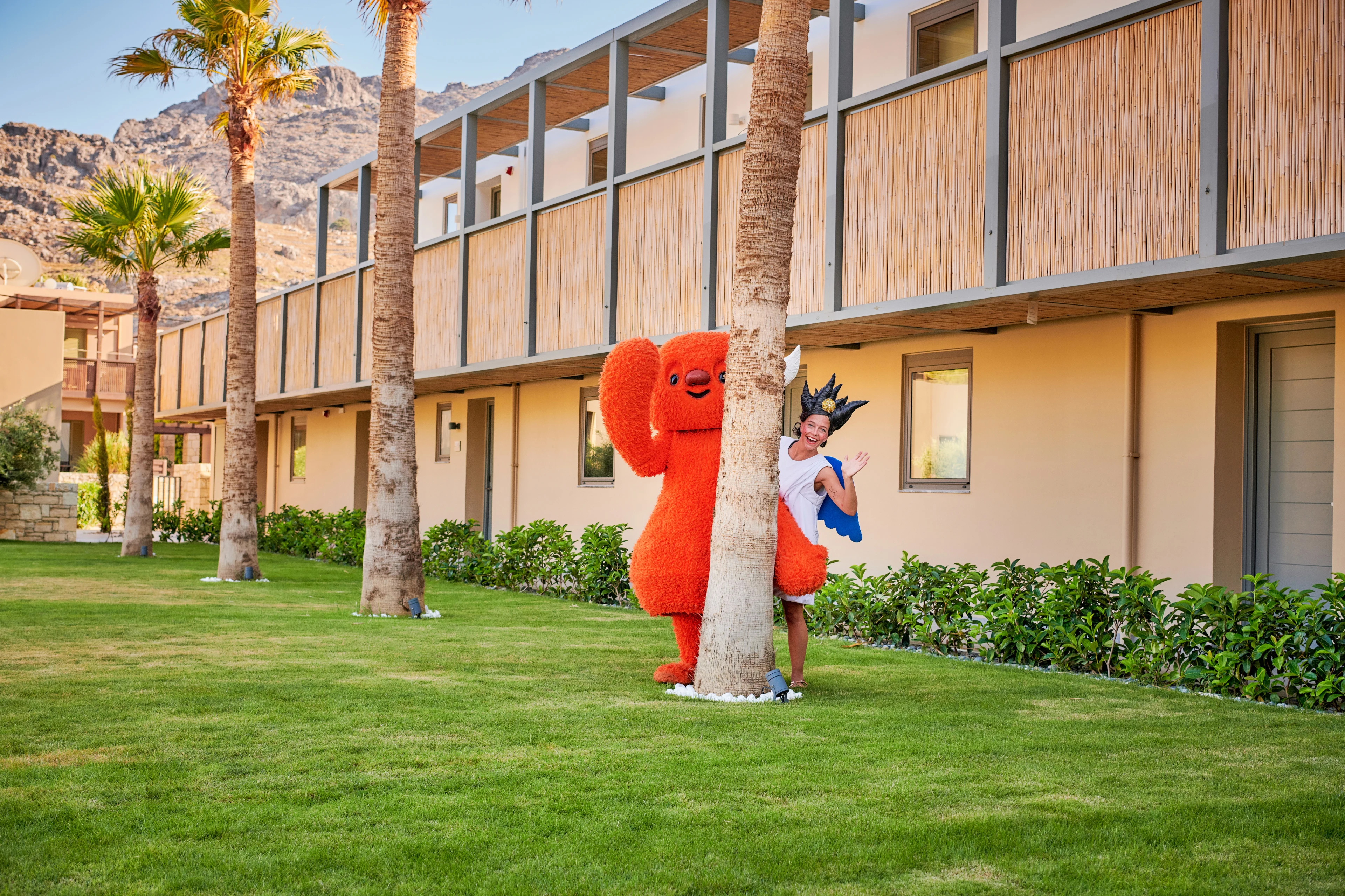 Polly & Mino står och vinkar bakom en palm på Kreta