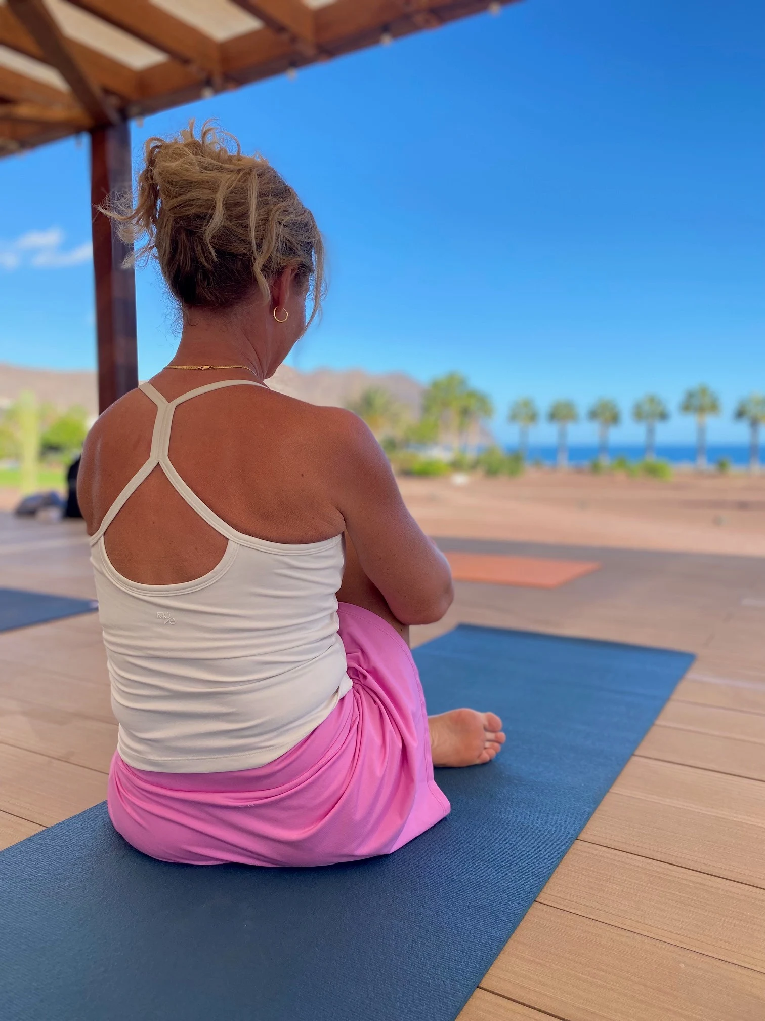 Dame som sitter med ryggen til på en blå yogamatte på et tredekke  ikledd hvit singel og rosa treningsskjørt. Blå himmel og palmer med havet som skimtes i horisonten.