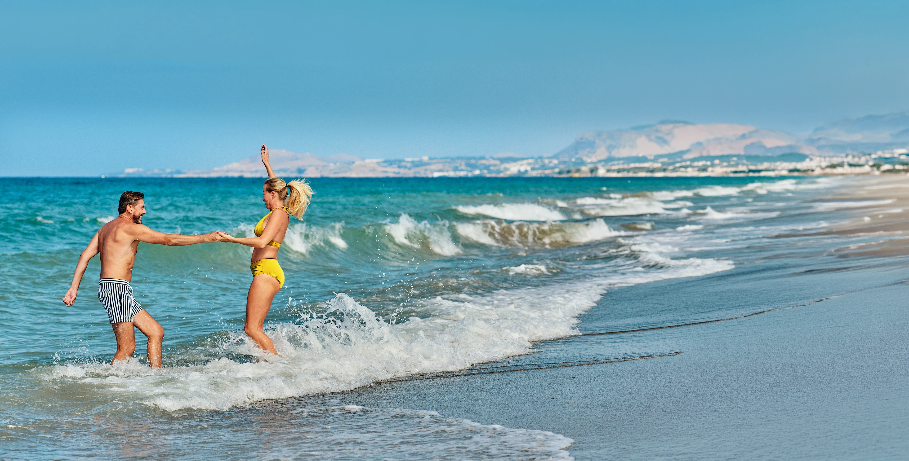 Två personer leker i vågorna vid en sandstrand med havet och kustlinjen i bakgrunden.
