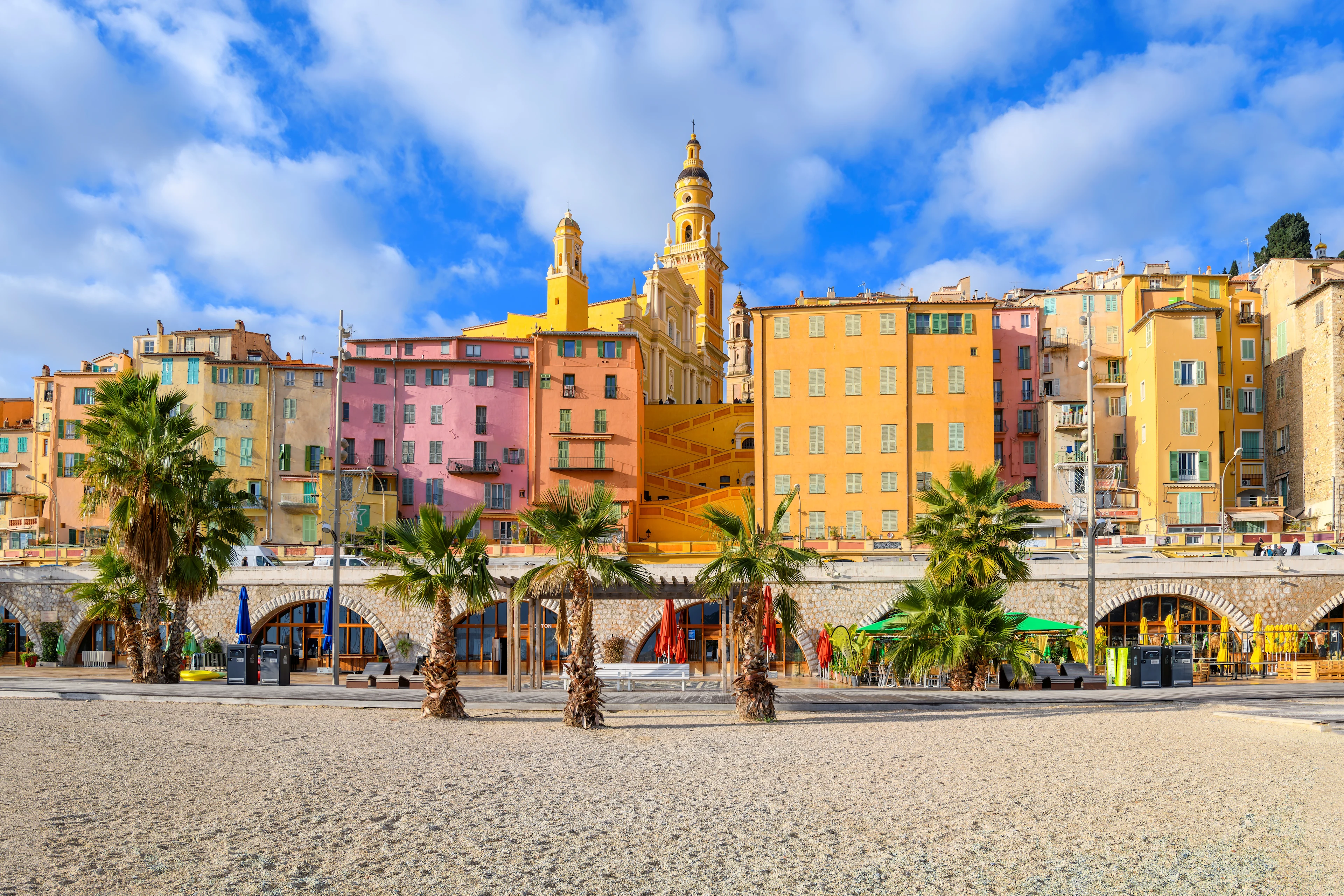 Färgglada hus och strandpromenaden vid Plage des Sablettes i Menton på Franska rivieran, med palmer och den historiska stadskärnan i bakgrunden.