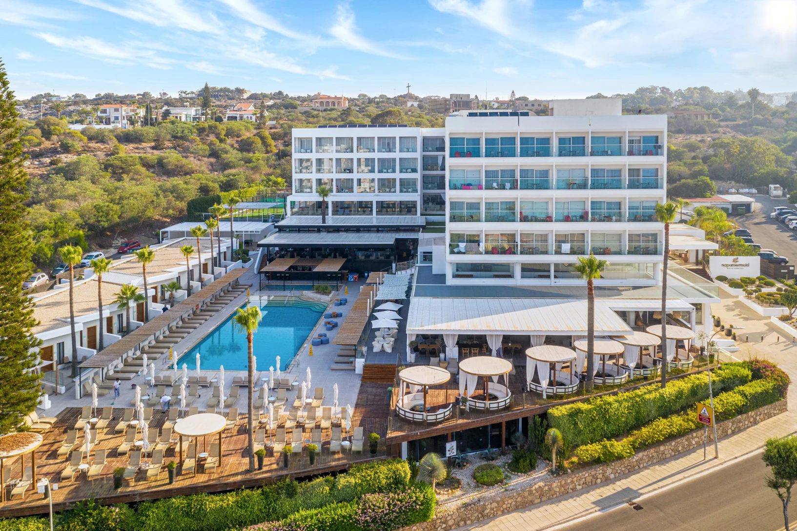 Ett stort modernt hotell med flera terrasser, solstolar och en utomhuspool, omgivet av grönska och beläget vid en väg.