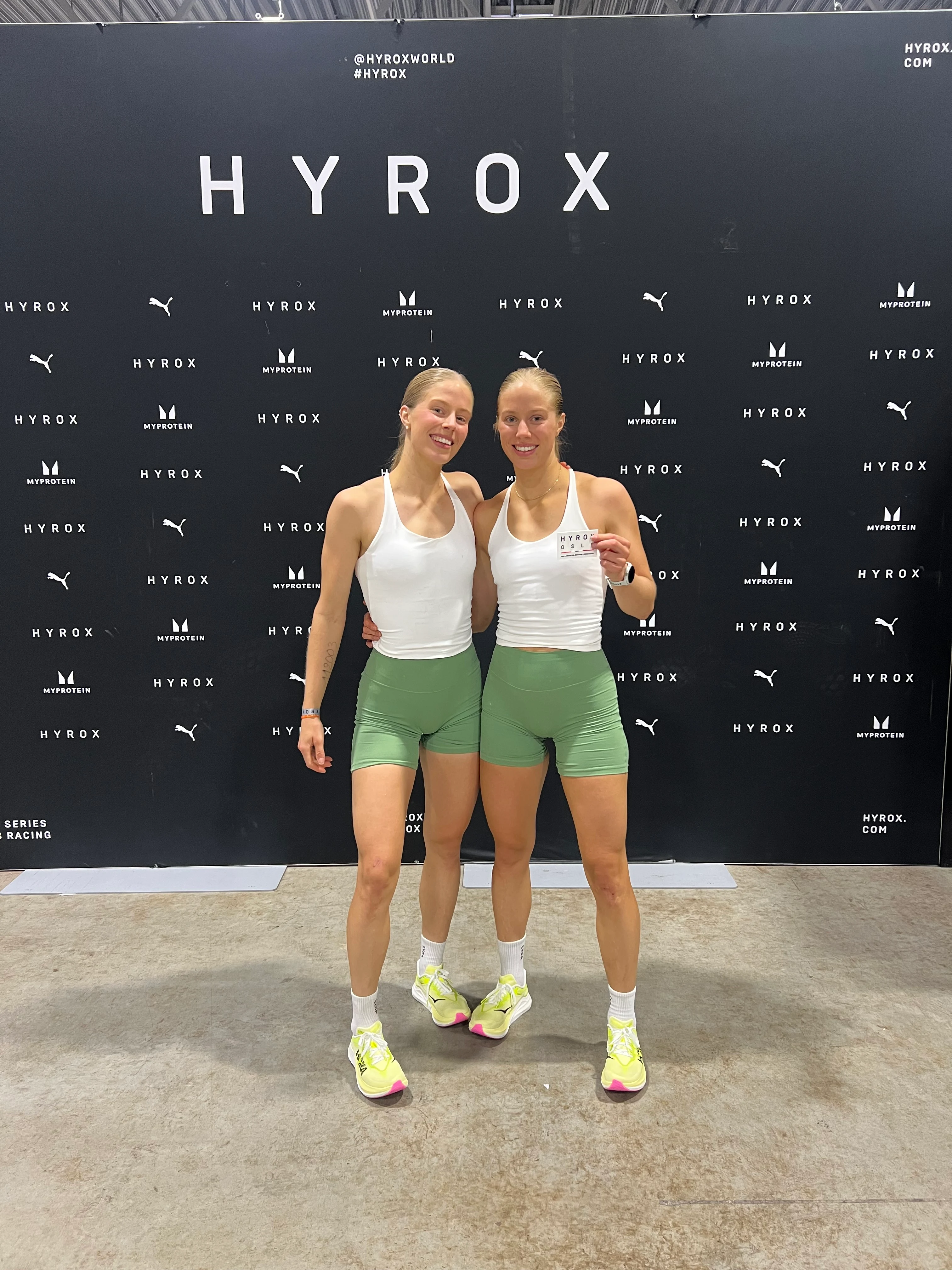 Elin og Guro kledd i hvite singleter og grønne treningsshorts står foran en sort vegg med hvit HYROX skrift