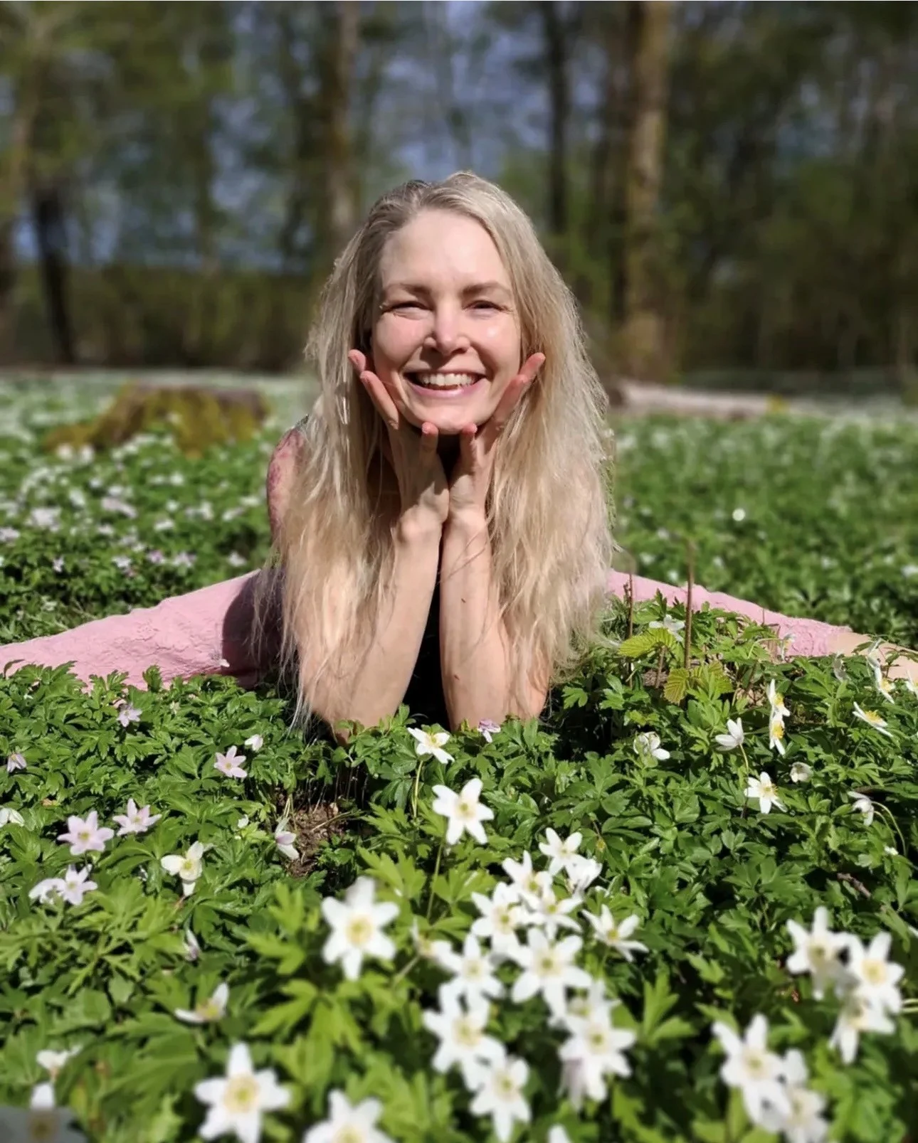 Person ligger bland vita vårblommor i en grön äng med skog i bakgrunden