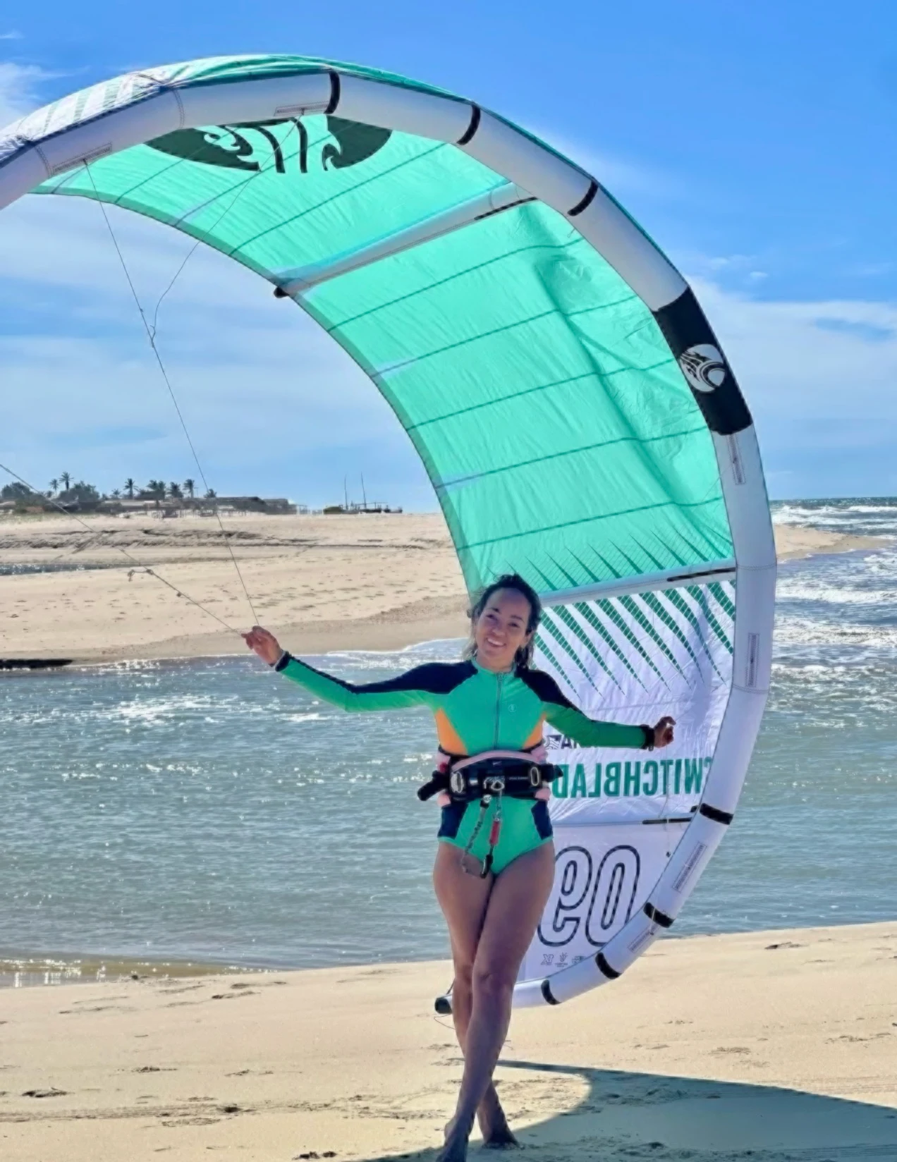 Profilbilde: Thatiana som står på en sandstrand i turkis våtdtakt og holder et turkist kiteseil i begge hender. 