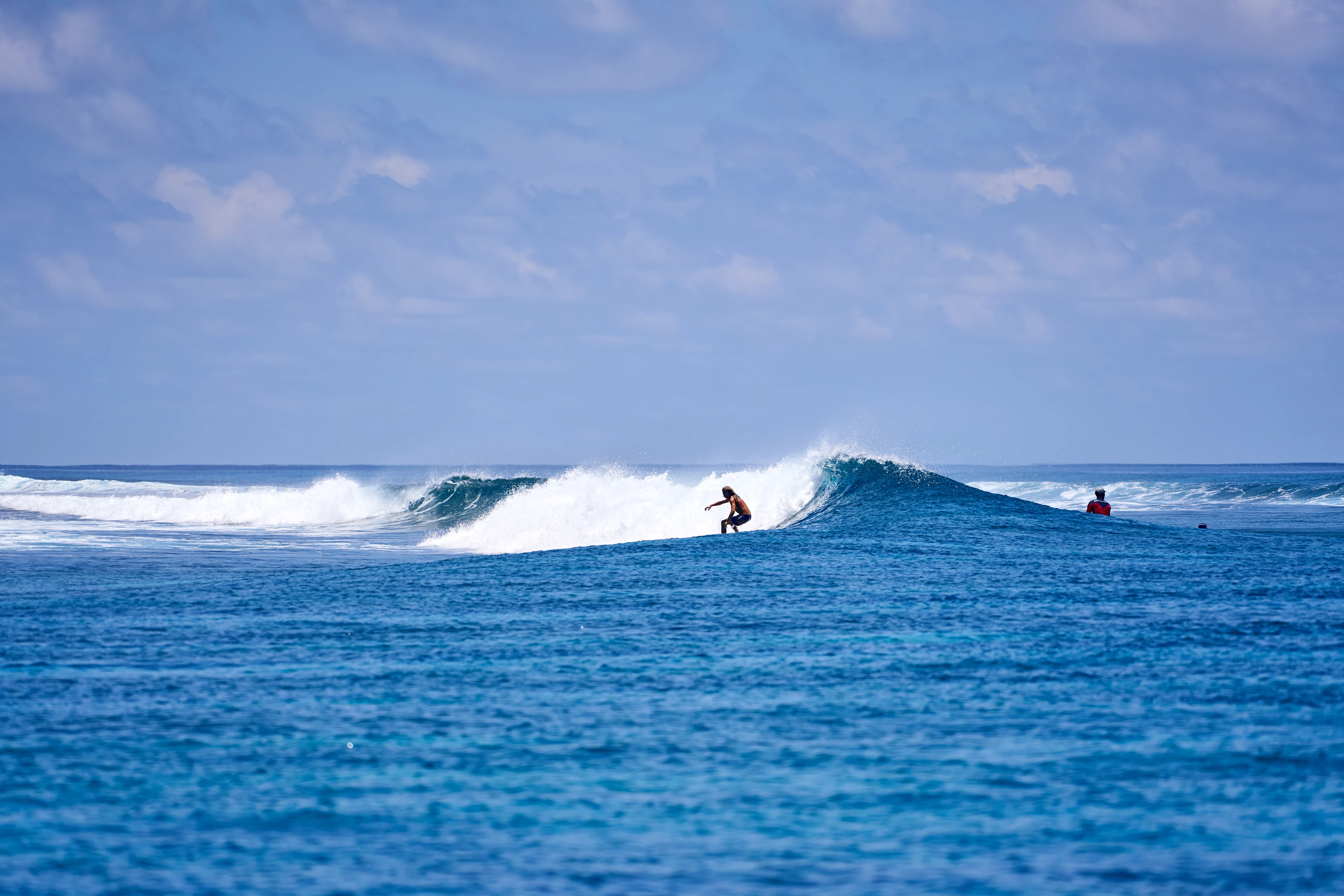 En person surfer på en bølge, mens en anden sidder i vandet i baggrunden