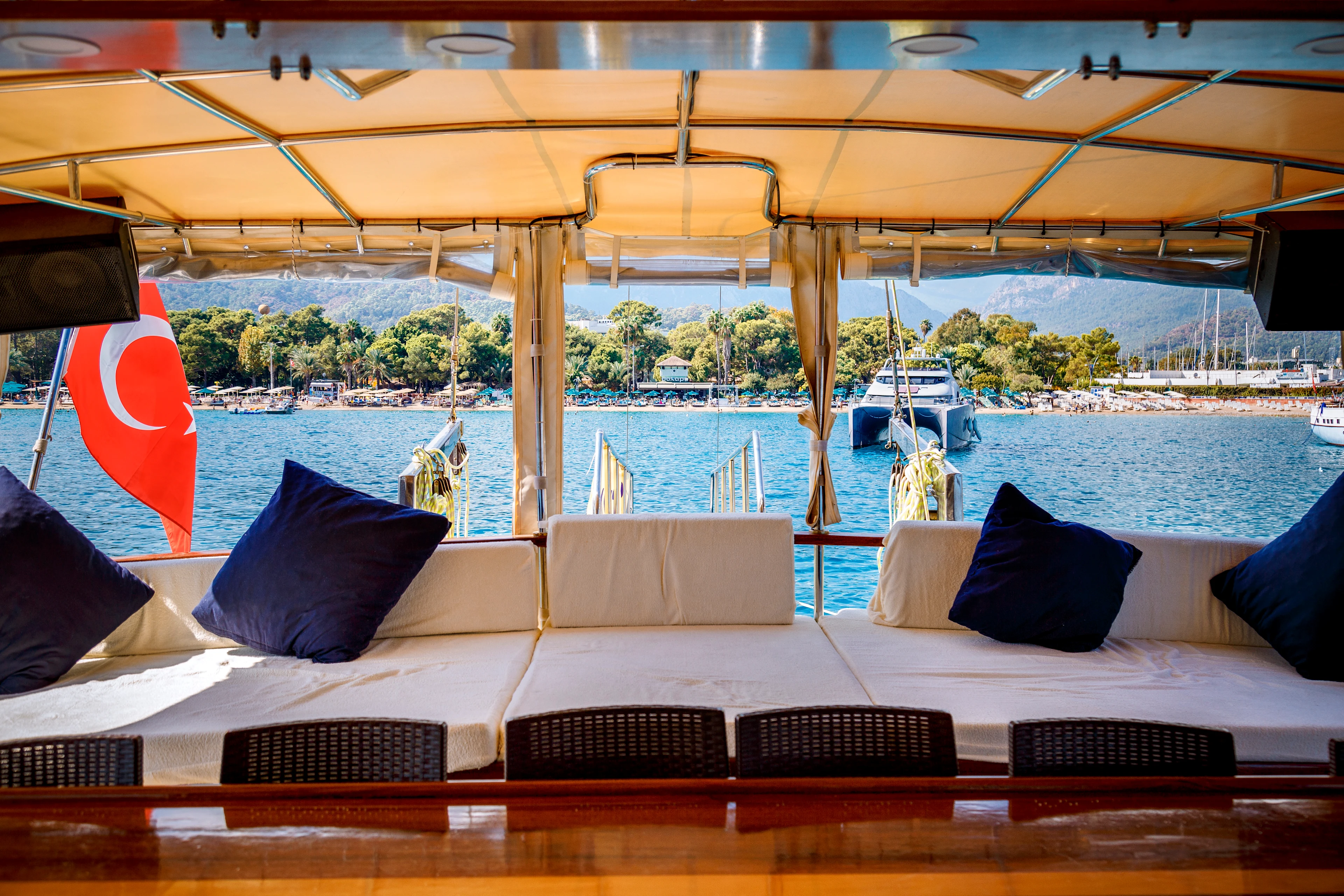 Näkymä veneestä, jossa on varjoisa oleskelutila satamaan päin ja taustalla metsäiset vuoret