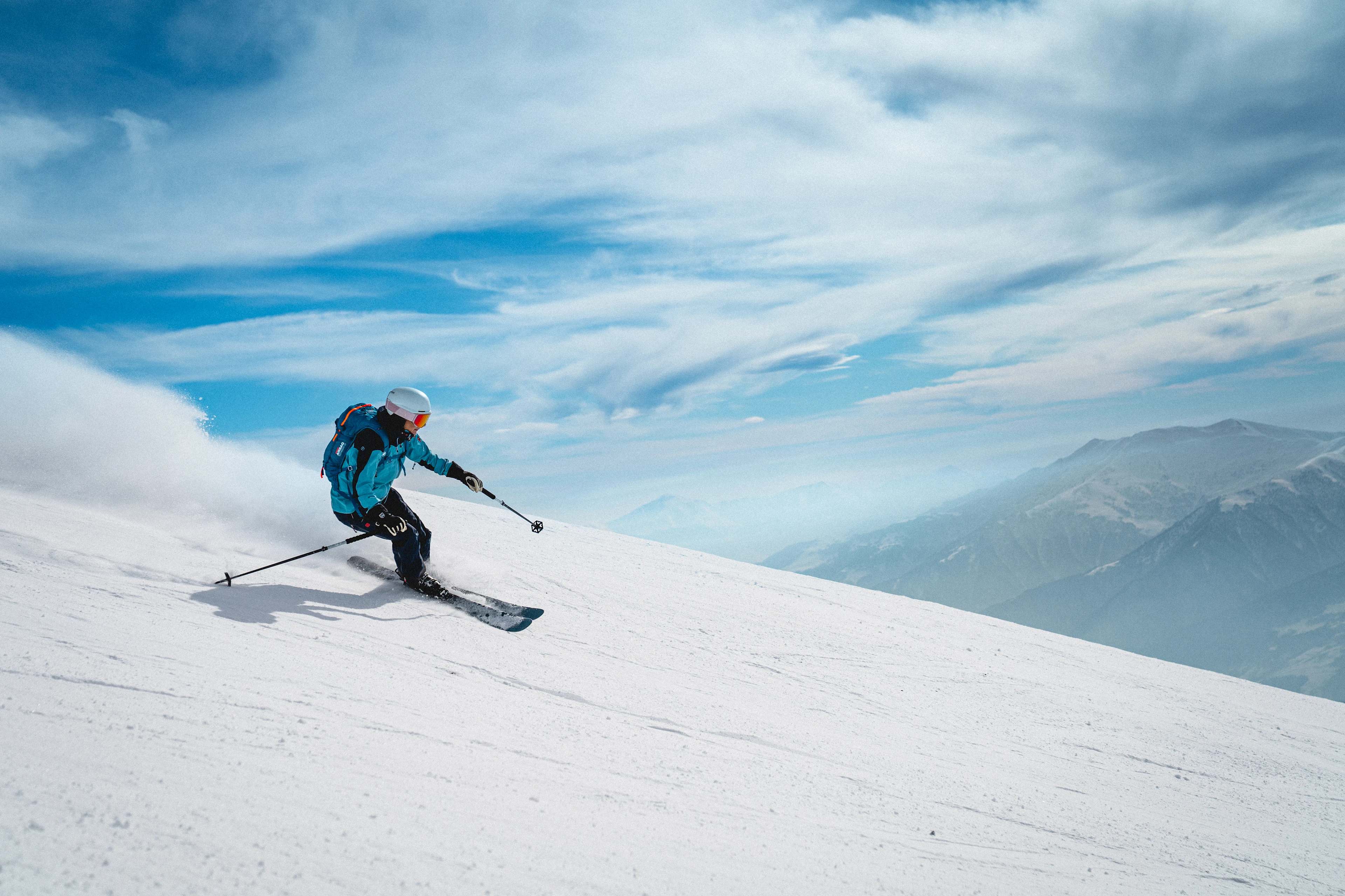 Skidåkare åker utför en snötäckt bergssida i Gudauri, Georgia, med vidsträckta berg och blå himmel i bakgrunden.