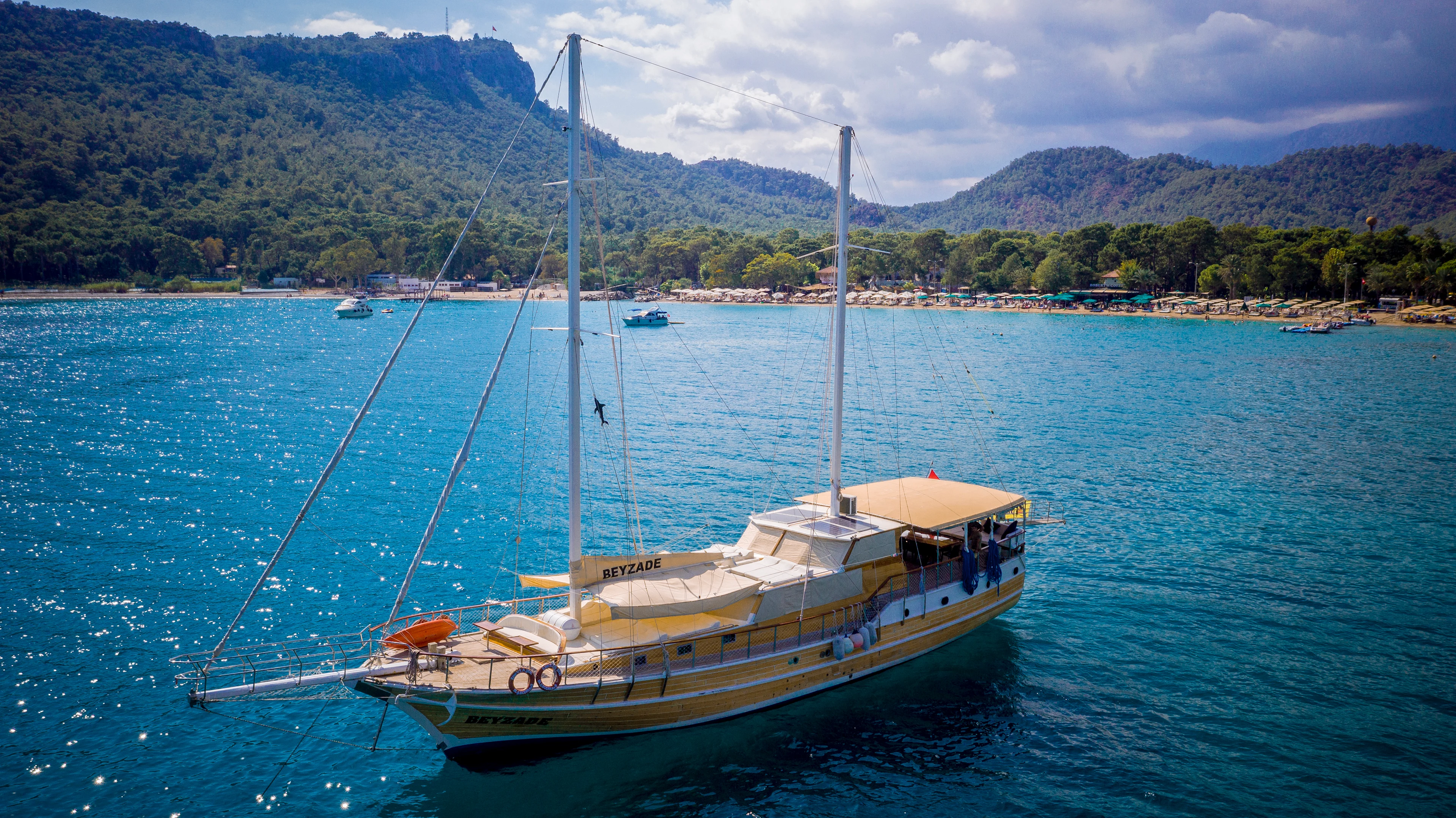 En träsegelbåt ligger i klart blått vatten nära en strand med skogsklädda berg i bakgrunden