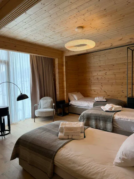 Lyst træhotelværelse med tre senge, store vinduer med gardiner og håndklæder pænt lagt ud på sengene