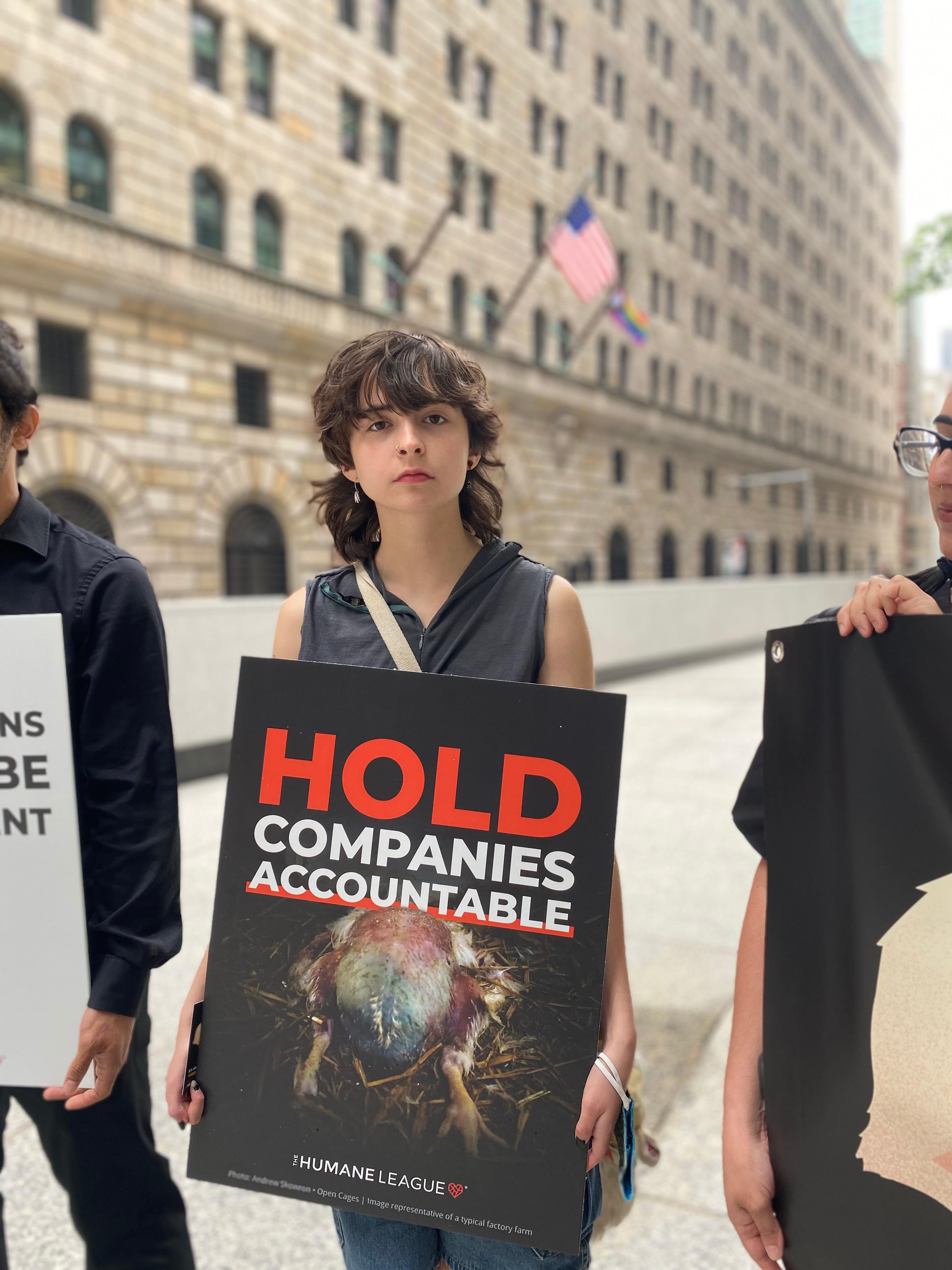 Protester holds a sign that reads: Hold corporations accountable, with an image of a factory farmed chicken raised for meat