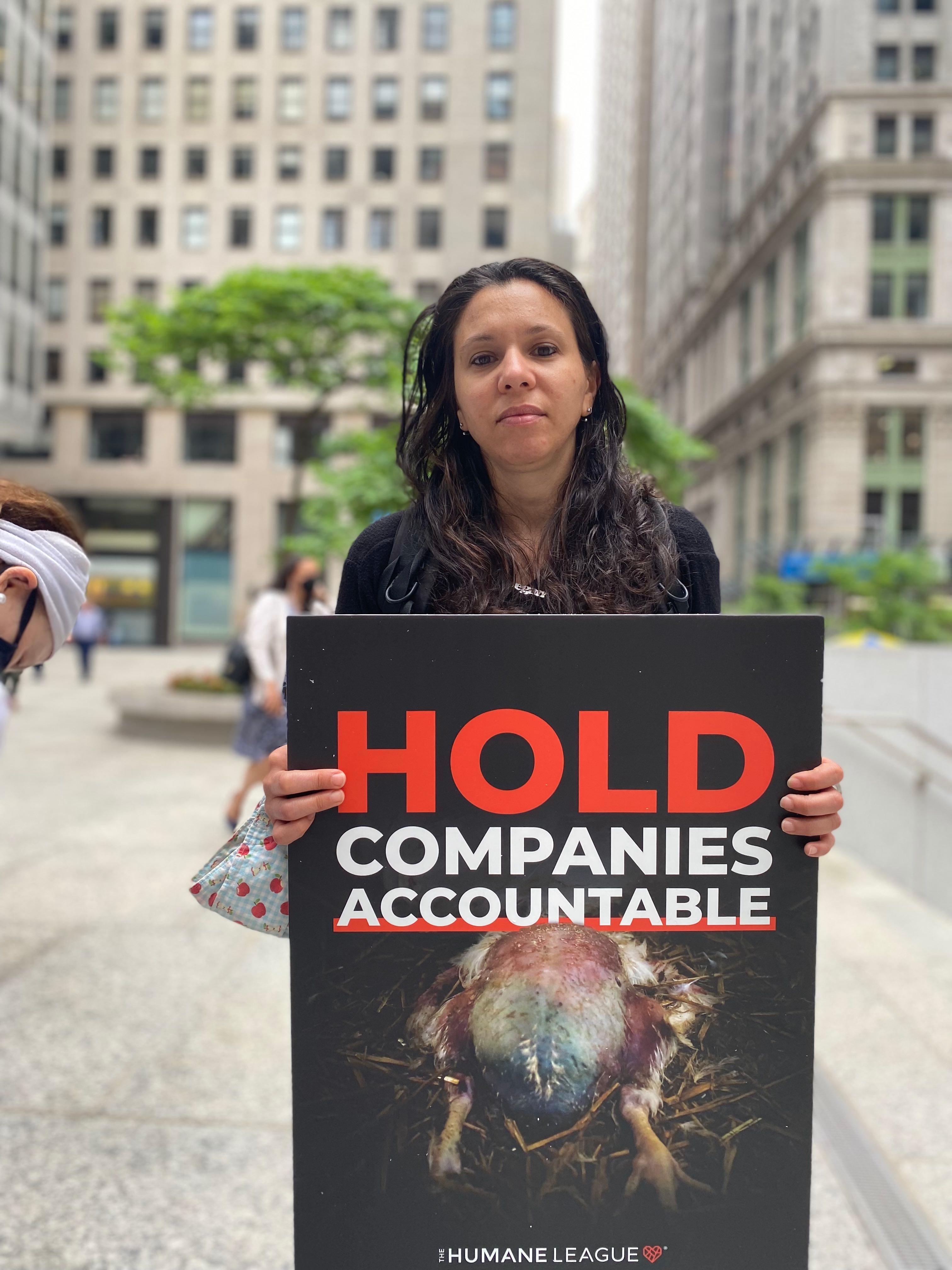 Protester holds a sign that reads: Hold corporations accountable, with an image of a factory farmed chicken raised for meat