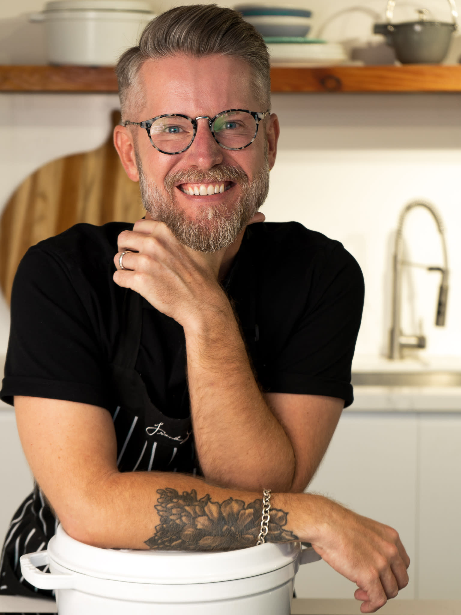 Chris Tucker smiles in his kitchen, wearing glasses and a black T-shirt. 