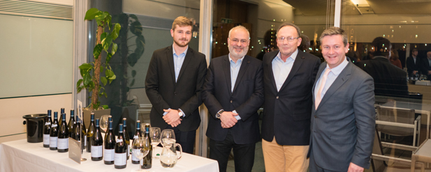 Four men in suits standing behind a table of wine